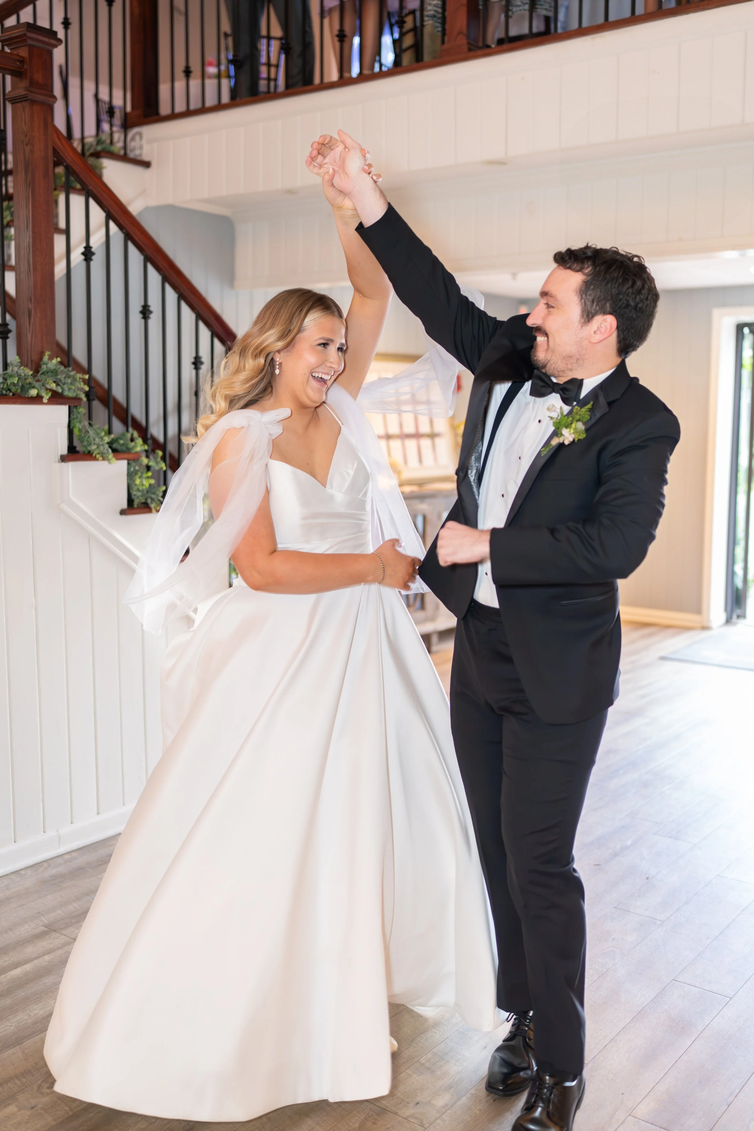 Happy bride and groom dancing indoors, holding hands, smiling, with a staircase and plants in the background.