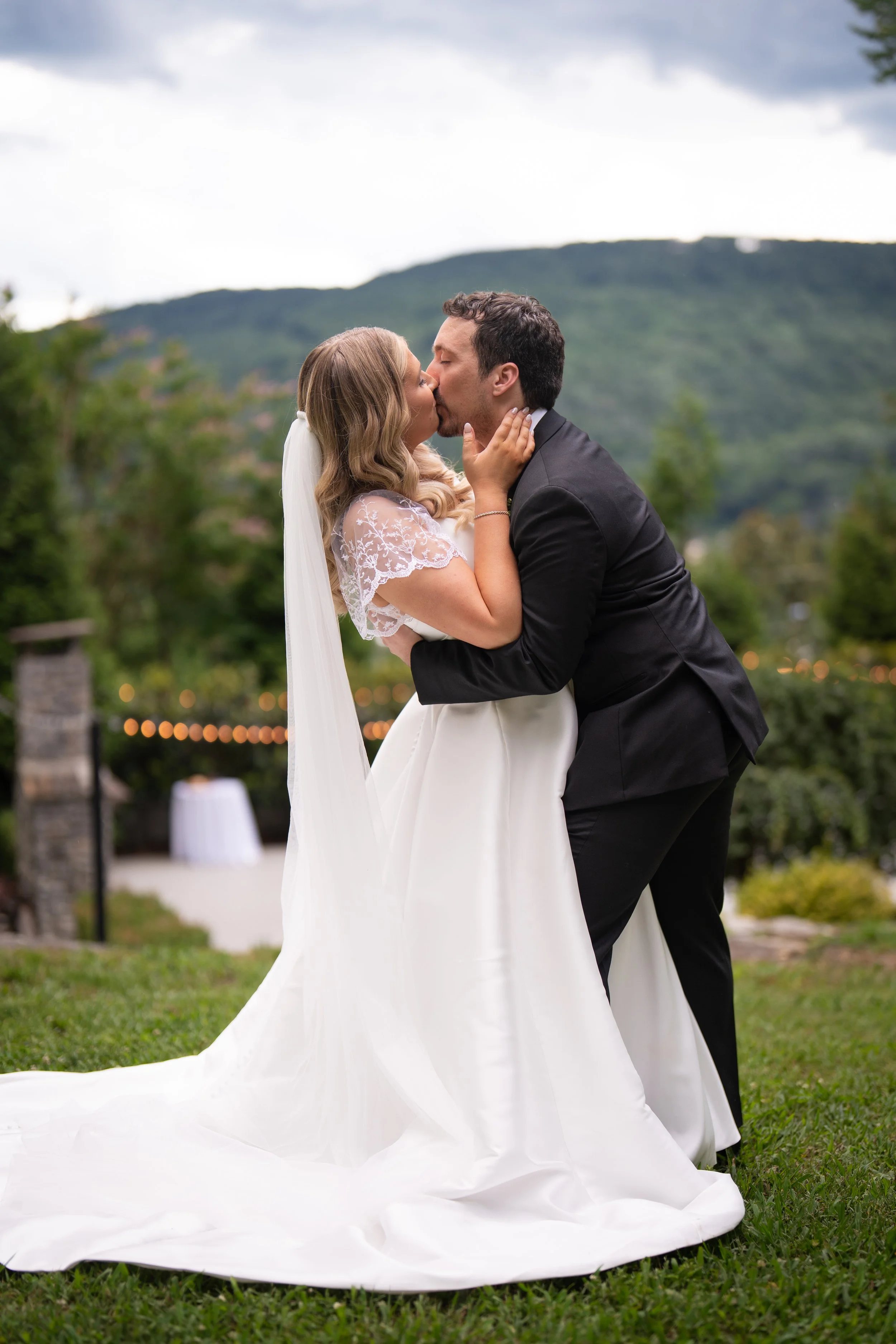 A bride and groom share a kiss outdoors during their wedding, with a scenic mountain backdrop and string lights.