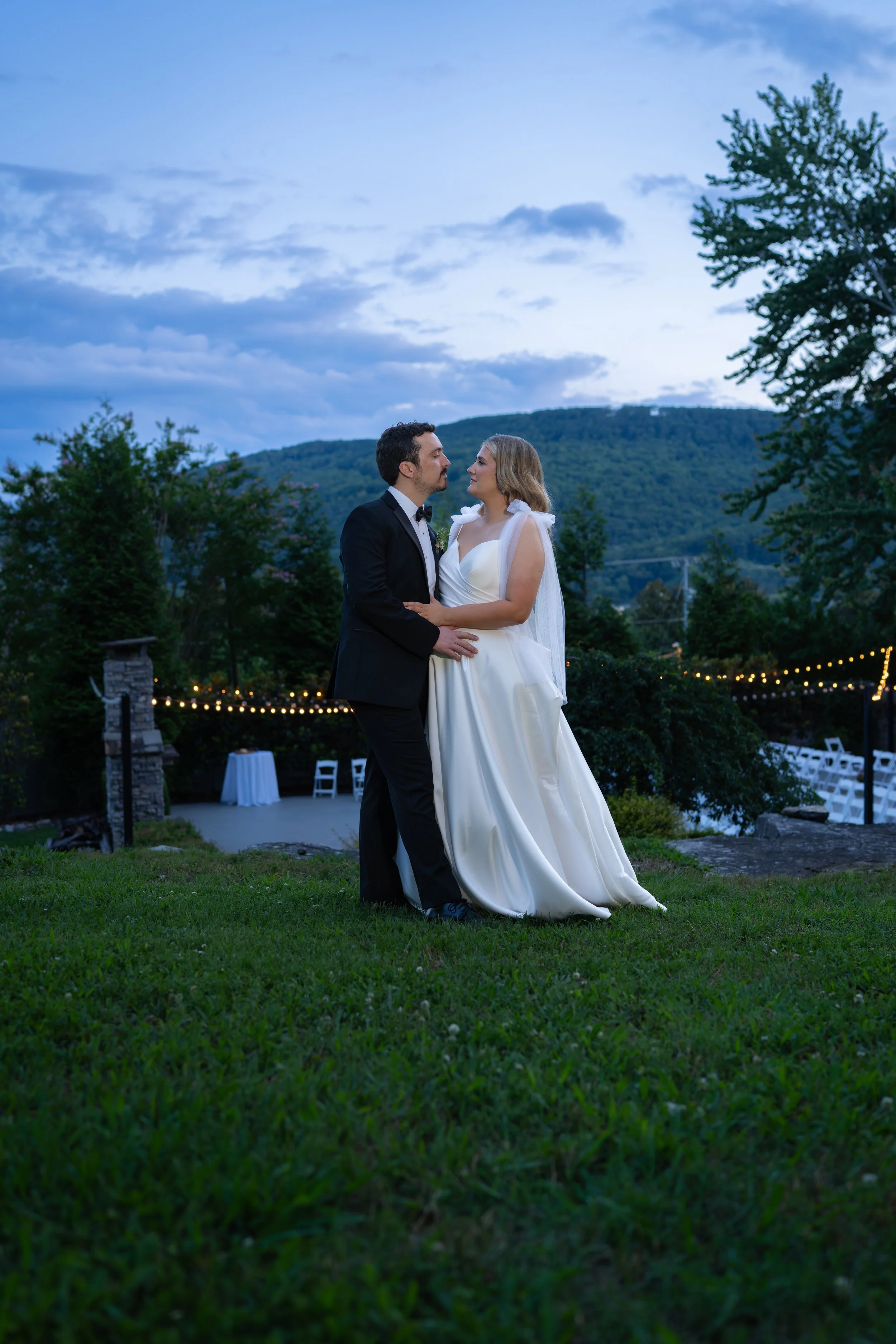 A bride and groom are dancing outdoors at dusk in a scenic setting with hills, trees, and string lights in the background.