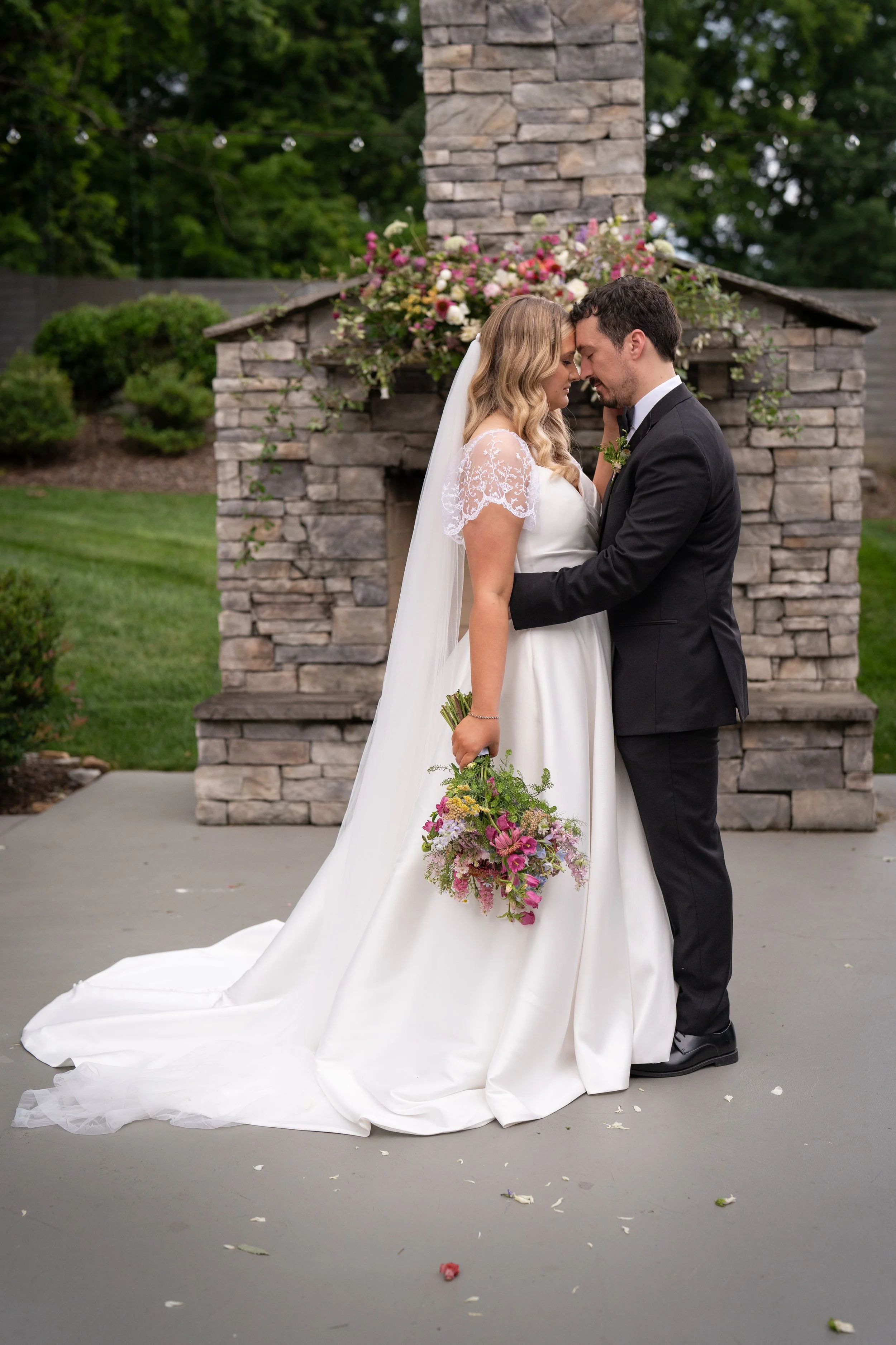 A bride and groom close together with foreheads touching, standing outdoors in front of a stone fireplace decorated with flowers, during their wedding.