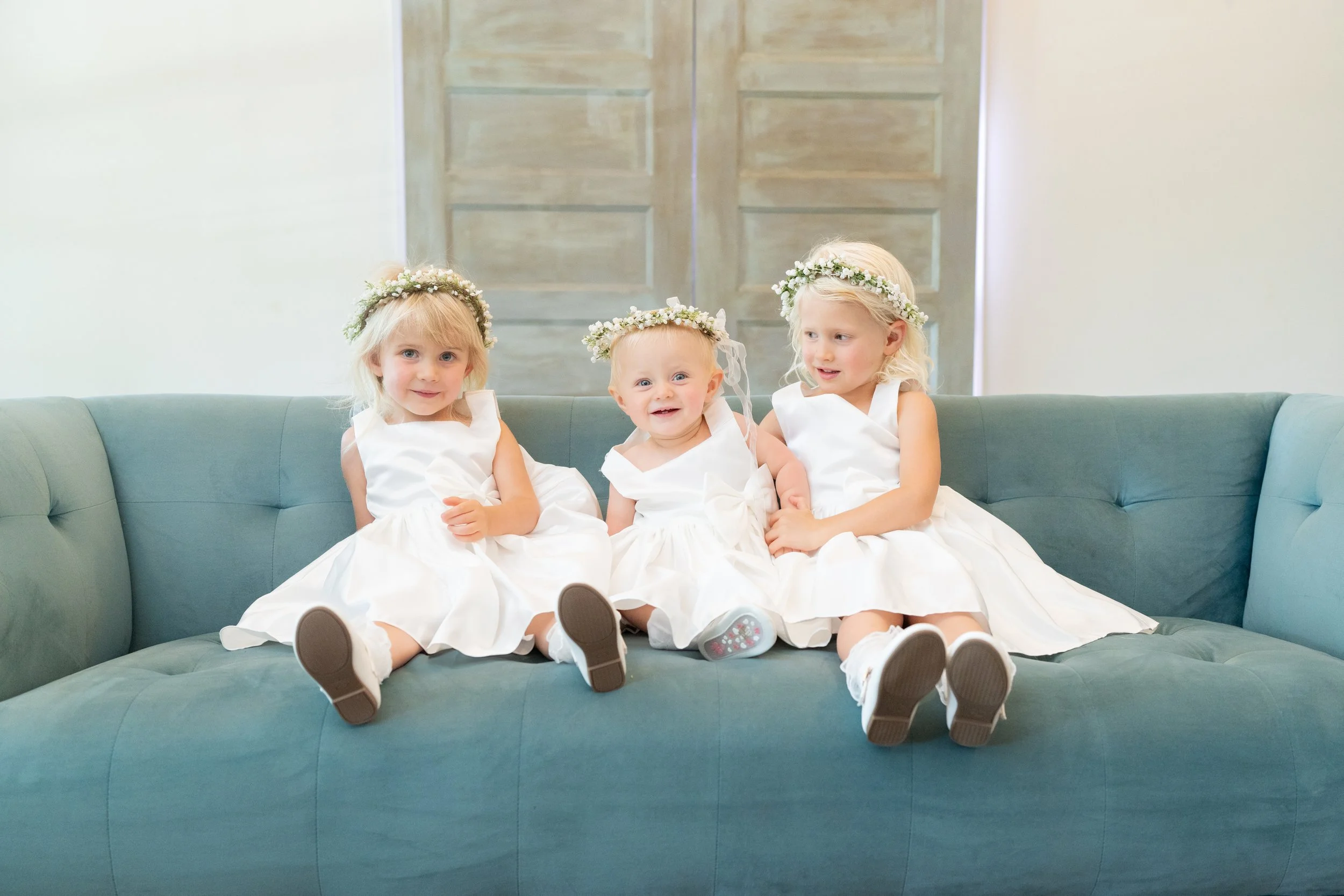 Wedding photos.   Three young girls sitting on a teal couch, wearing white dresses and floral crowns, during a special occasion.