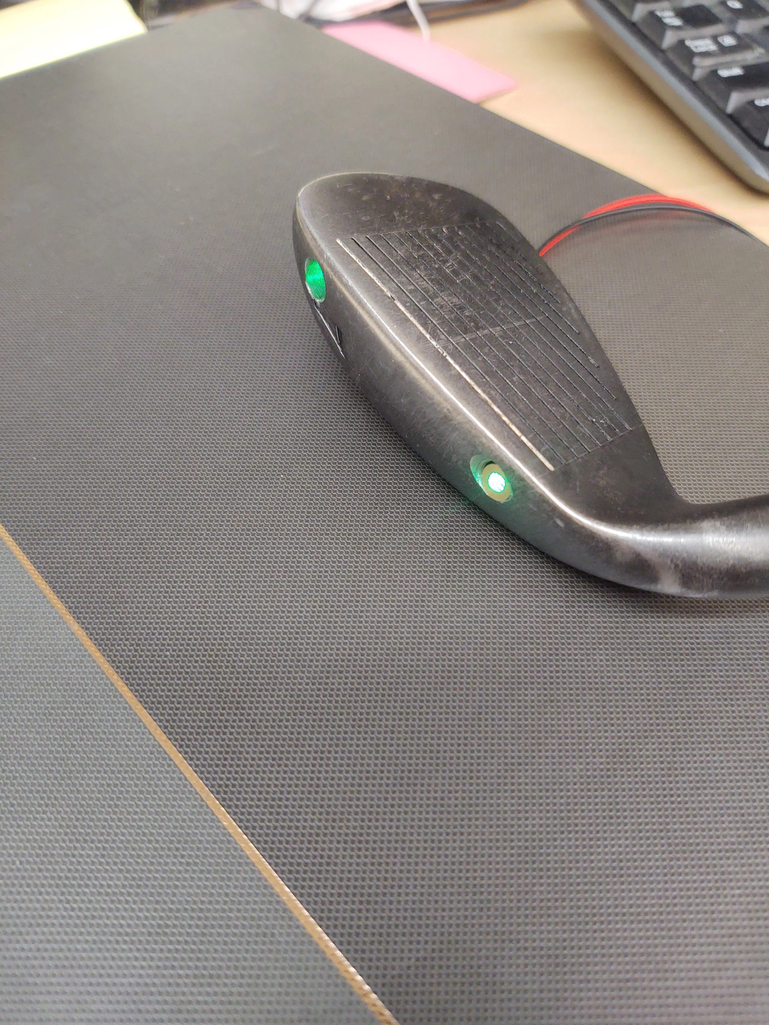 A golf club lying on a textured black mat with two green indicator lights on the clubhead. A computer keyboard is partially visible in the background.