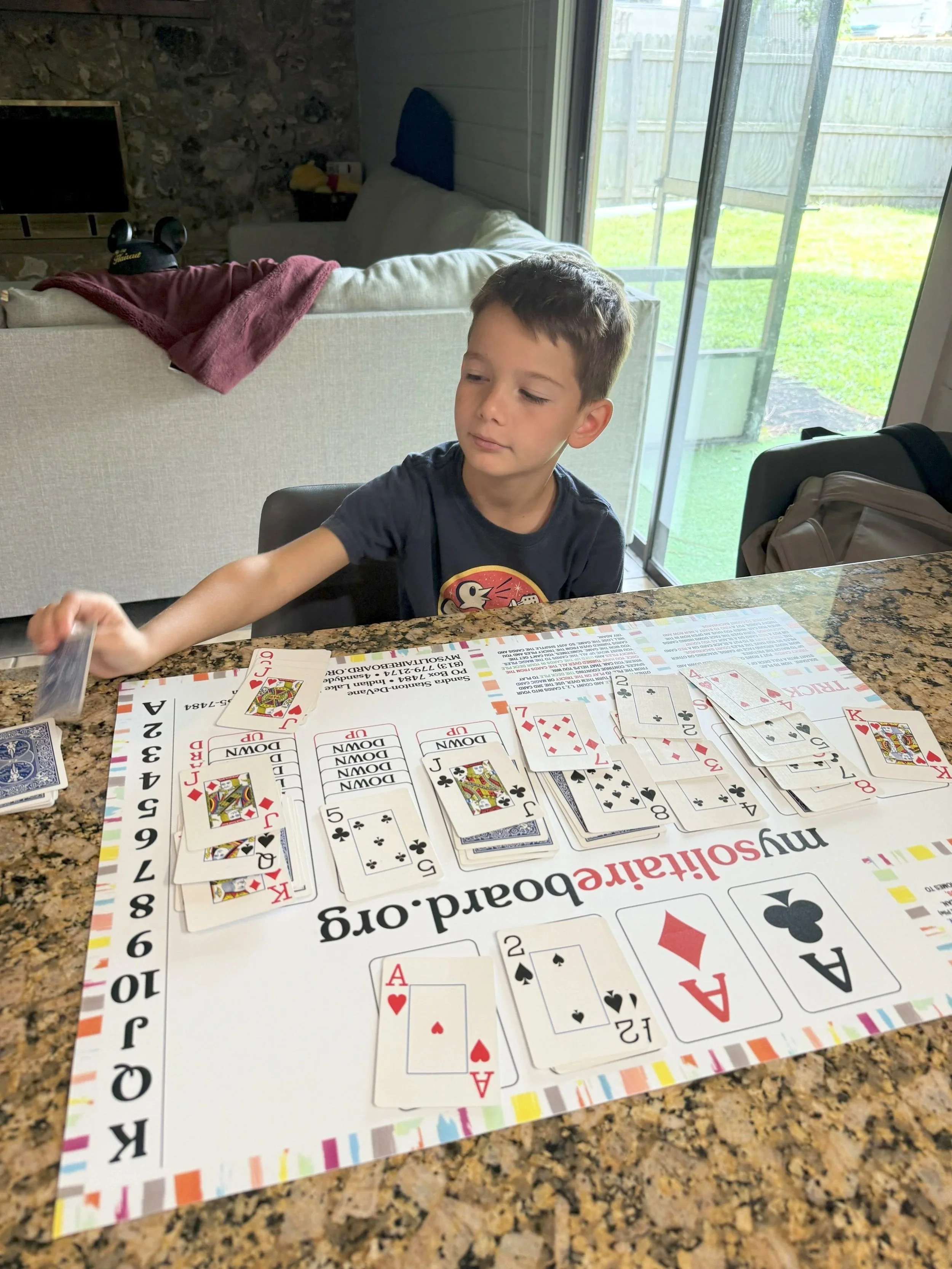 A young boy sitting at a kitchen table playing Janie's Game, an accessible Solitaire board designed for individuals with Down syndrome, autism, seniors, and caregivers.The table has a colorful solitaire mat, and the boy is reaching to pick up a card.