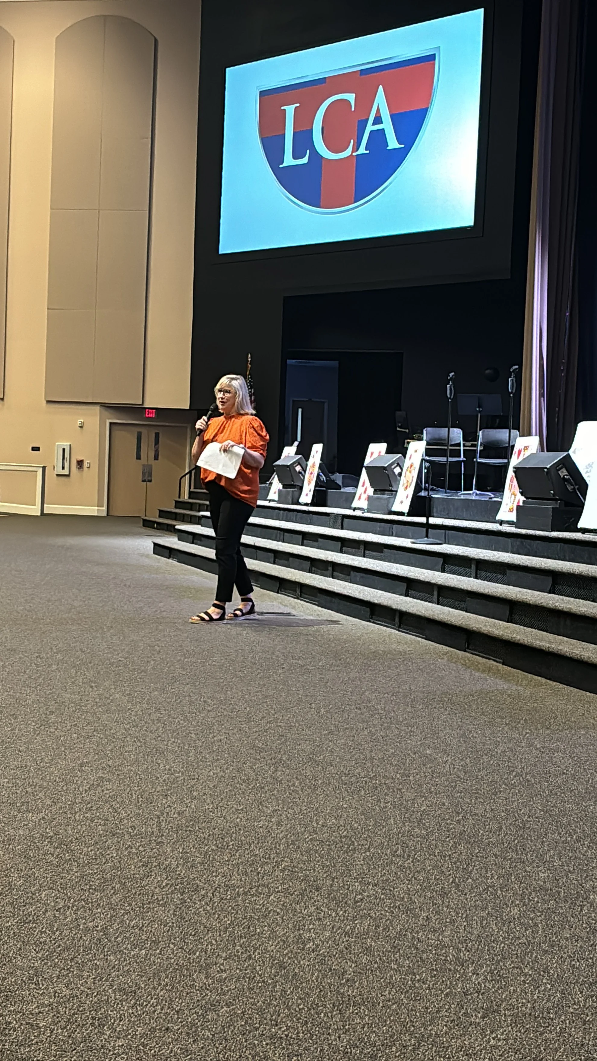 A woman with white hair, wearing a bright orange top, black pants, and sandals, is standing on a stage holding a microphone and a piece of paper in front of a large screen displaying a logo with the text LCA, which resembles a road sign, and appears to be speaking to an audience.