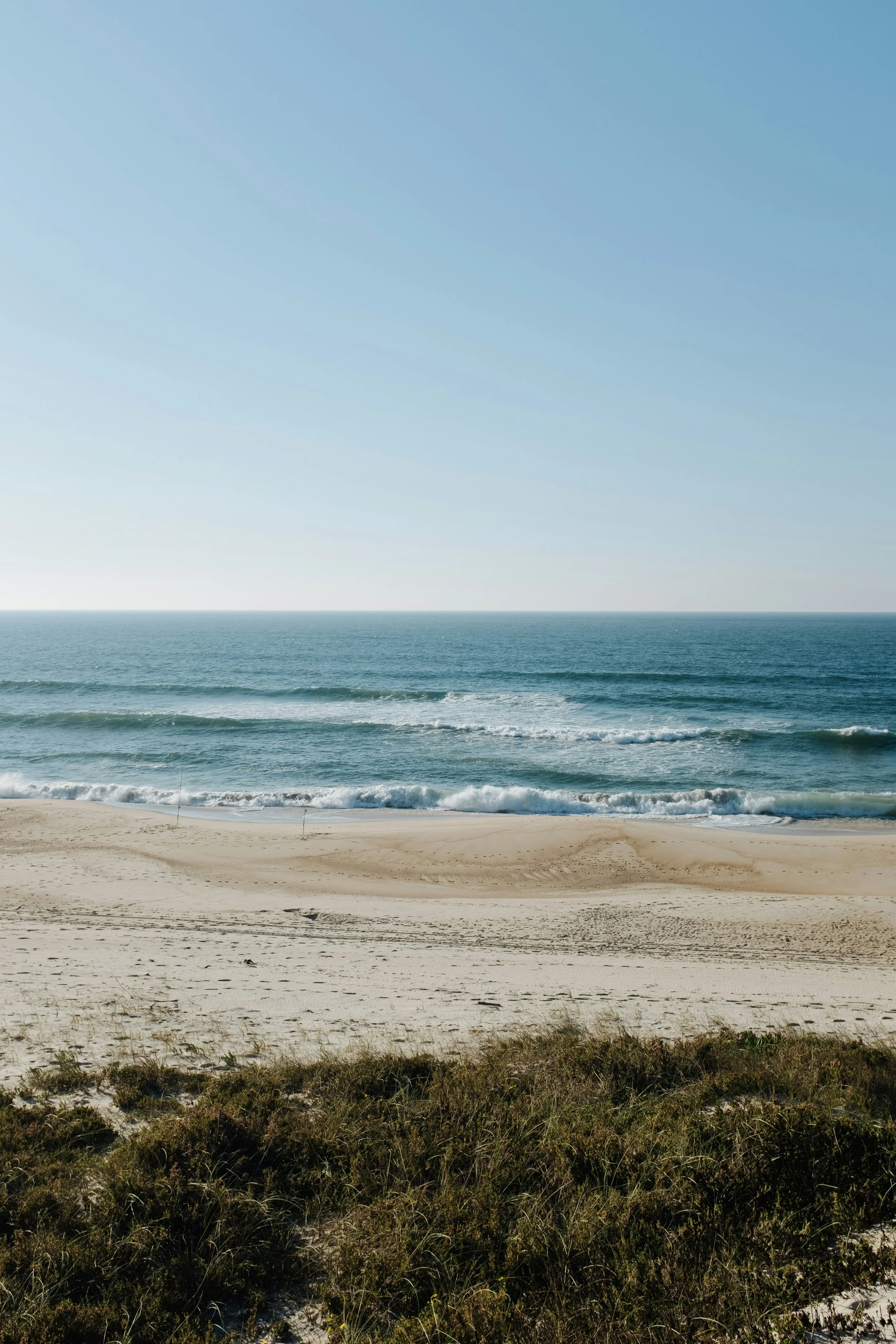 A sandy beach with some grass in the foreground, calm ocean waves, and a clear blue sky.