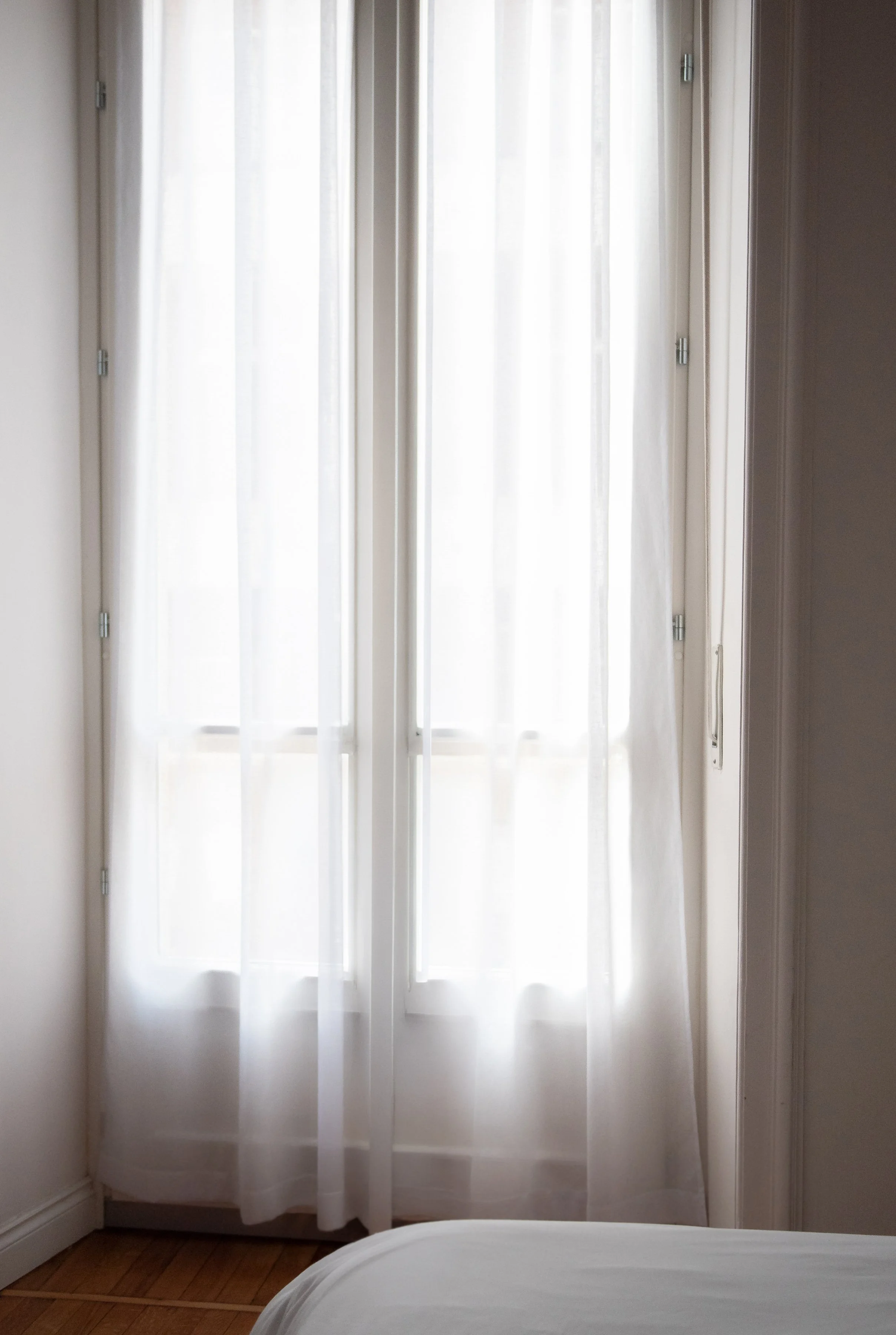 A white curtain covering a glass door with a metal handle, allowing light to softly filter into the room. Part of a bed with white sheets is visible at the bottom of the image.