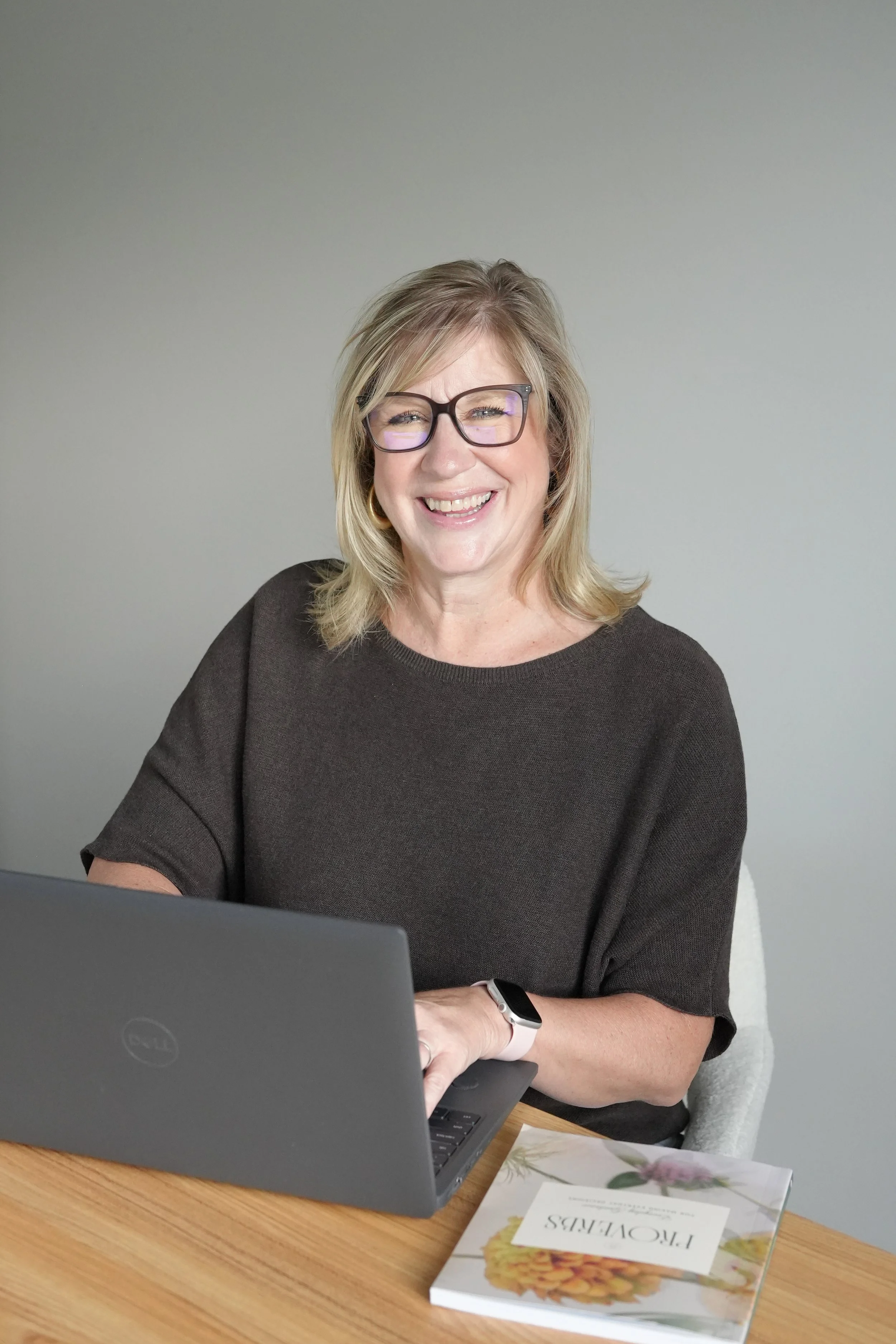 A woman with blonde hair, glasses, and a black top is smiling while sitting at a wooden table with a laptop and a floral book.