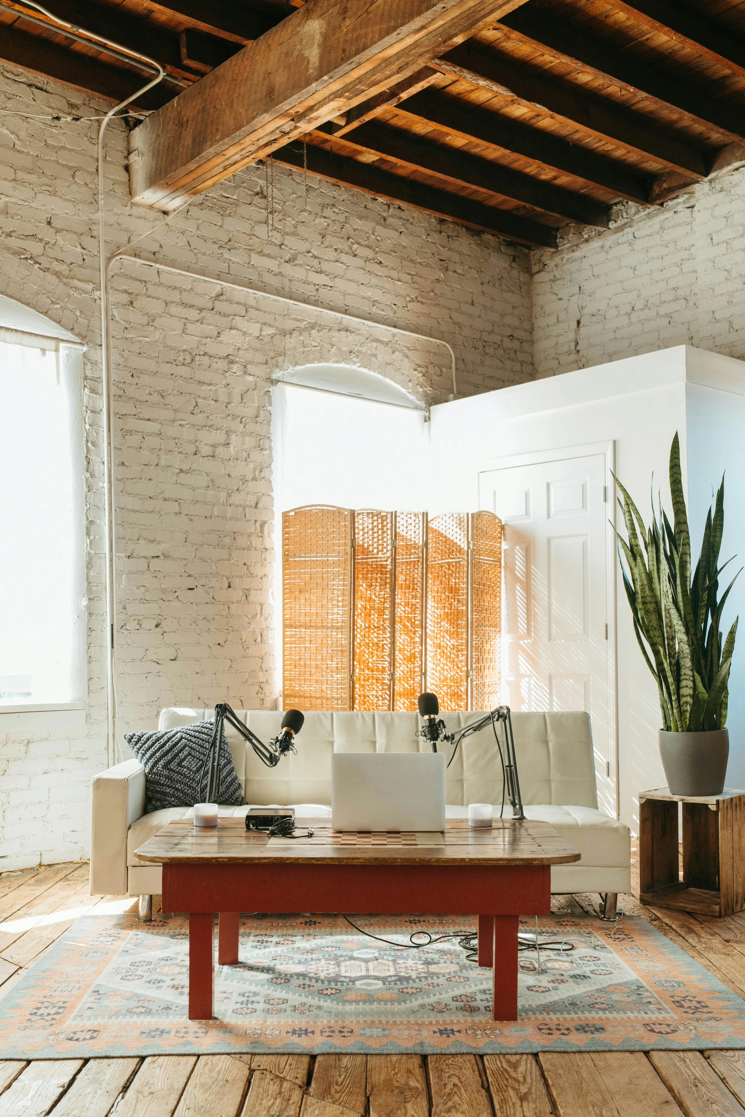 Interior of a bright room with white brick walls and wooden floors, featuring a white sofa with pillows, a wooden coffee table with a laptop and microphones, a plant in a large pot, and a decorative woven room divider, illuminated by natural light from windows.