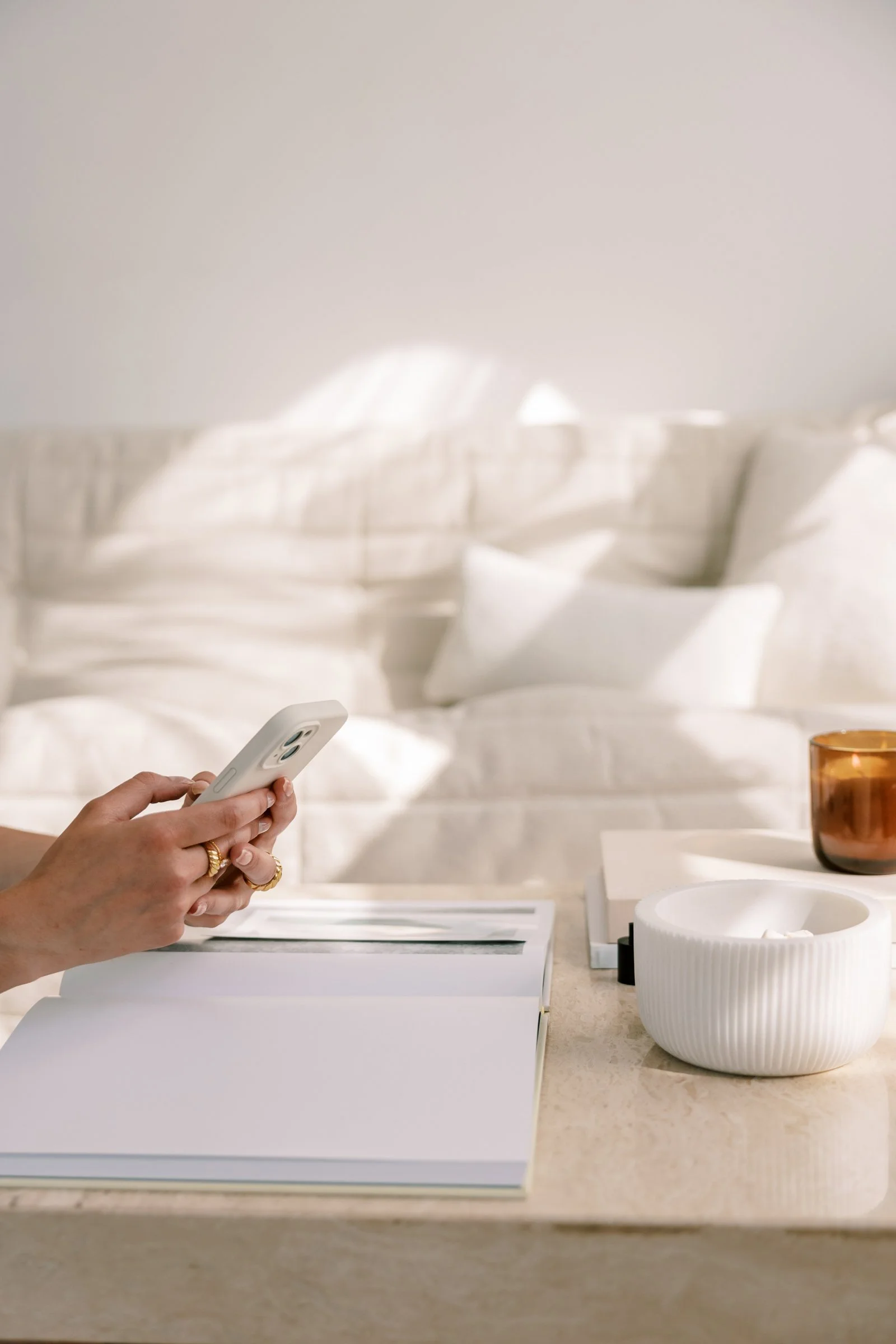 A person holding a white smartphone in a home setting, with a white bed and a bedside table holding a glass candle holder on the background.