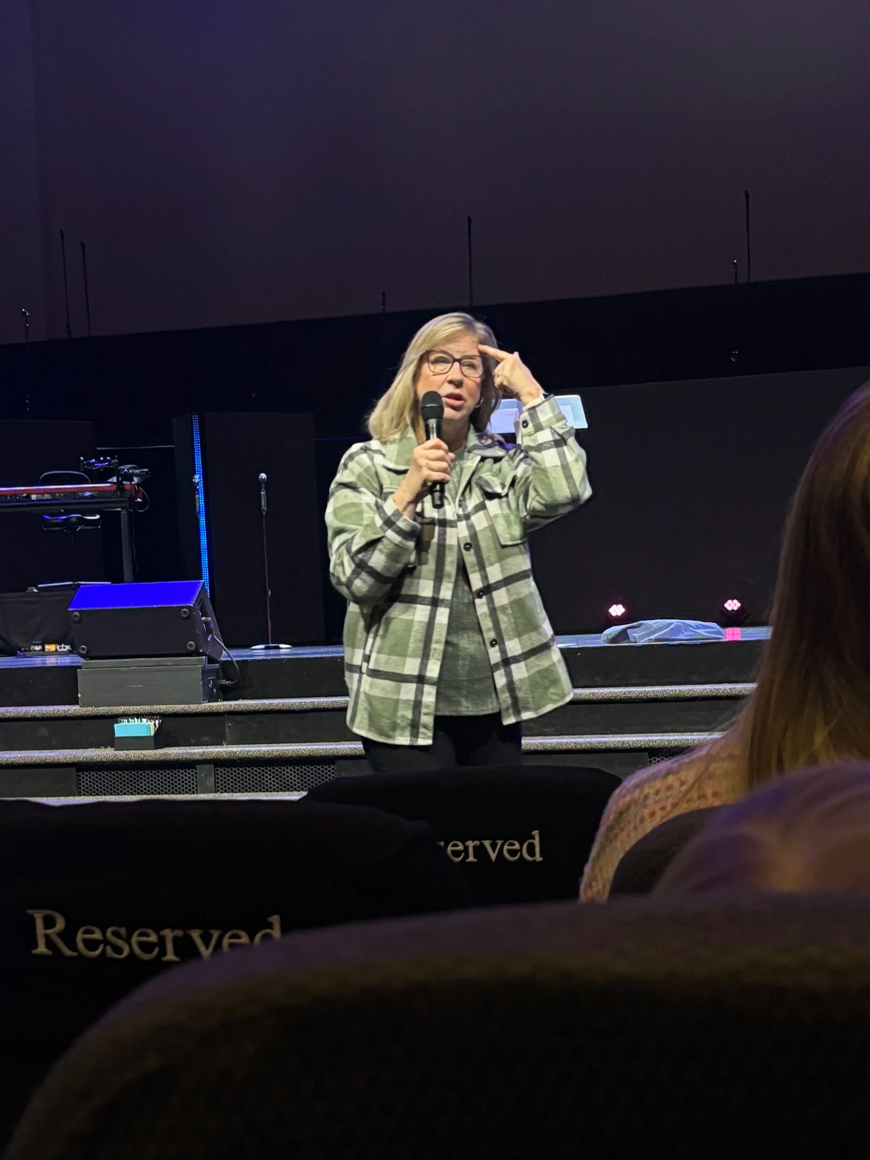Terrie Tollerson is standing on stage, holding a microphone and gesturing to her head with her index finger. She appears to be speaking to an audience in a dark room, with a large black screen behind her.