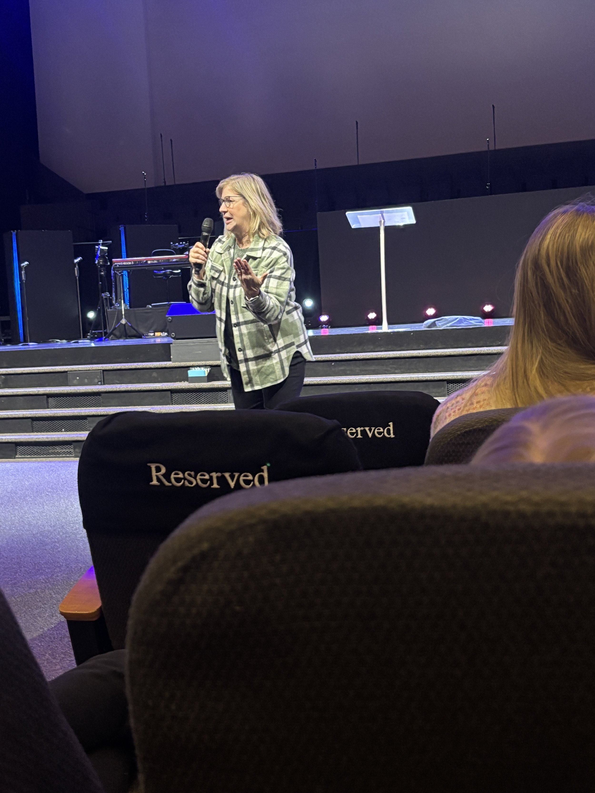 A woman with glasses and blonde hair is speaking into a microphone on stage. She is wearing a green and white plaid blazer. There are musical instruments and equipment in the background. The photo is taken from the audience, with the top of reserved theater seats visible in the foreground.