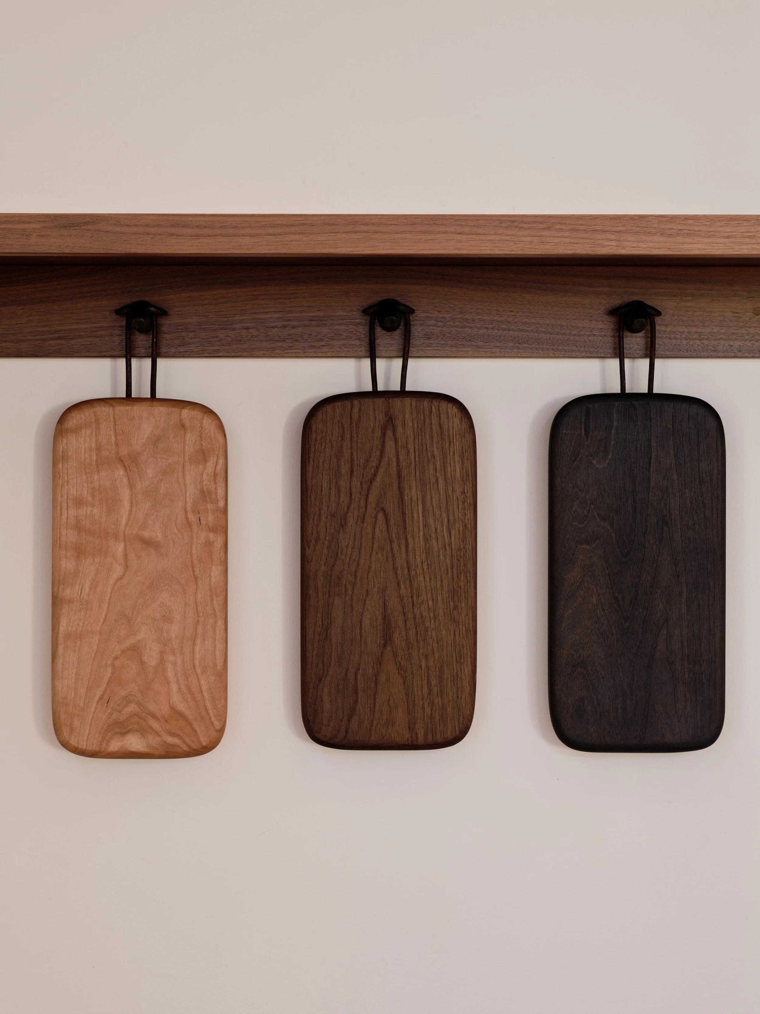 Three rectangular wooden cutting boards hanging on a wall hooks, from left to right: light, medium, and dark wood, against a plain background.