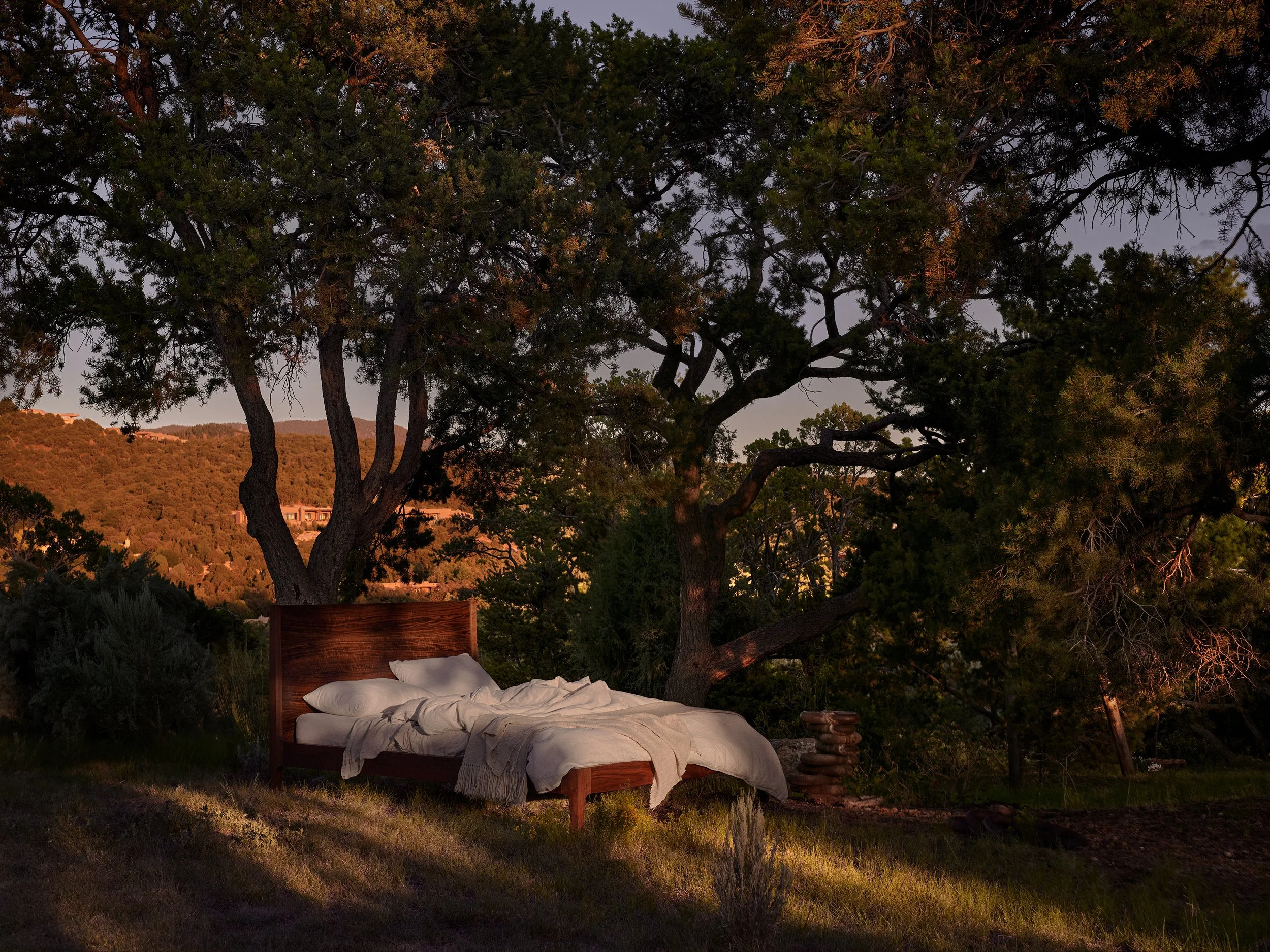 An outdoor bed with white bedding and pillows under a large tree, during sunset with warm lighting on the landscape.