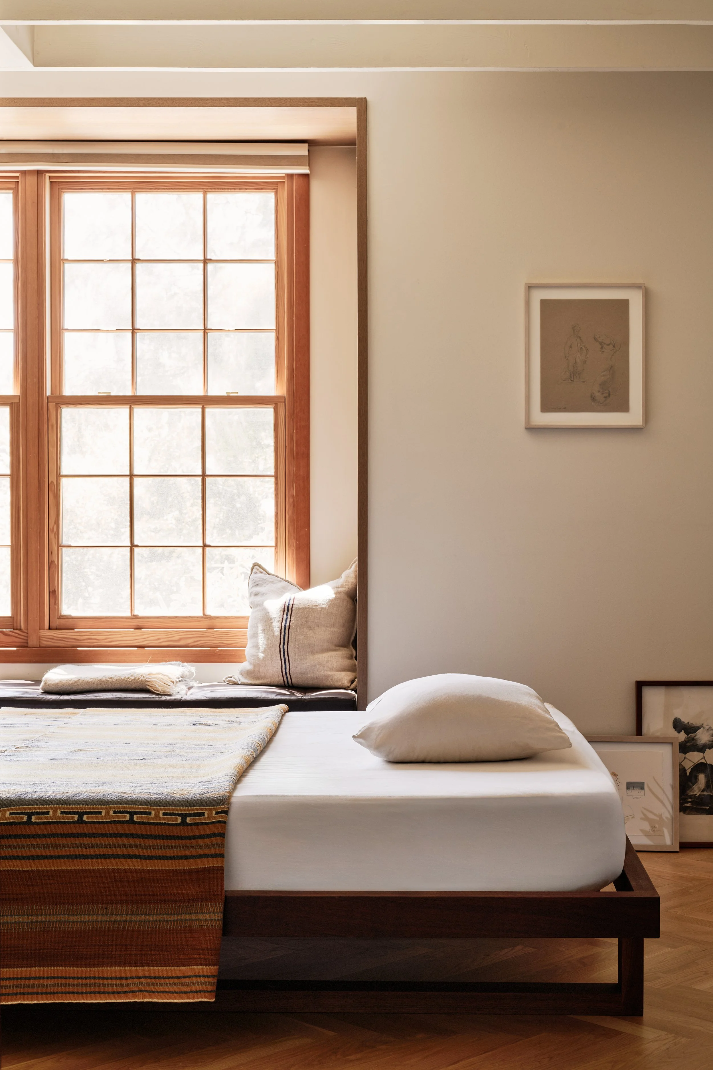 A bedroom with a wooden framed window, a bed with white sheets, a beige pillow, and a striped blanket. Decor items include framed artwork leaning against the wall and pillows by the window.