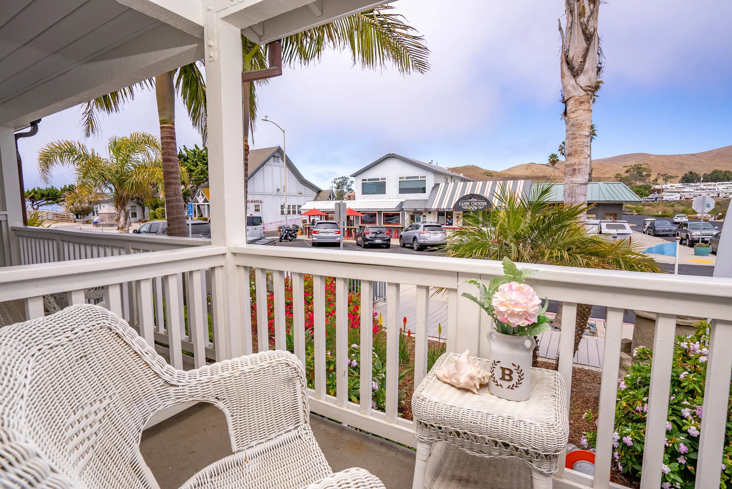 View from a porch overlooking a street with parked cars, palm trees, and a small commercial area with a shop and buildings. There's a white wicker chair and a small table with a pink flower arrangement and a shell on the porch.