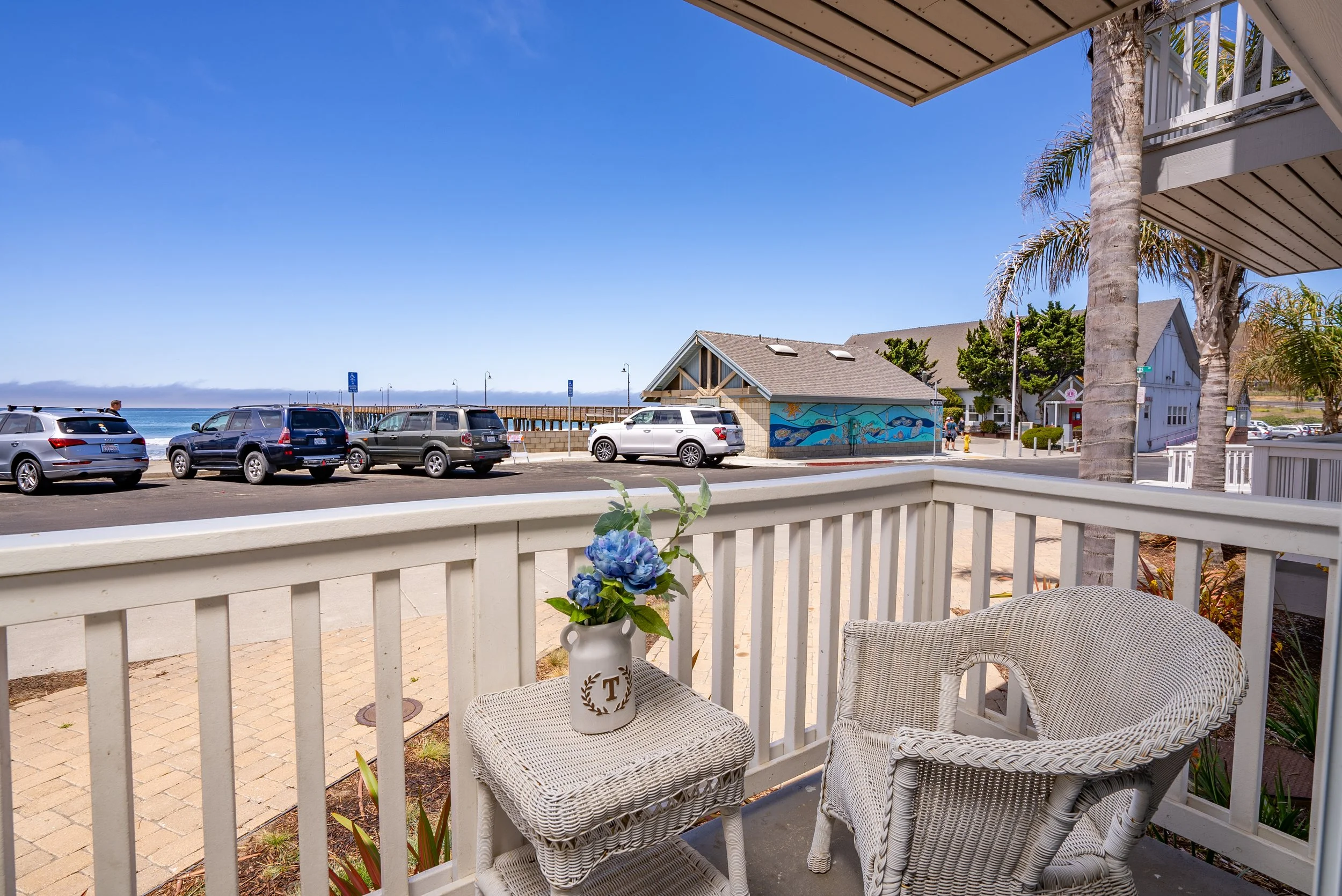 View from a porch overlooking a parking lot next to the ocean with several parked cars, a building with a mural, and palm trees, under a clear blue sky. There's a white wicker chair and a small table with a vase of blue flowers on the porch.