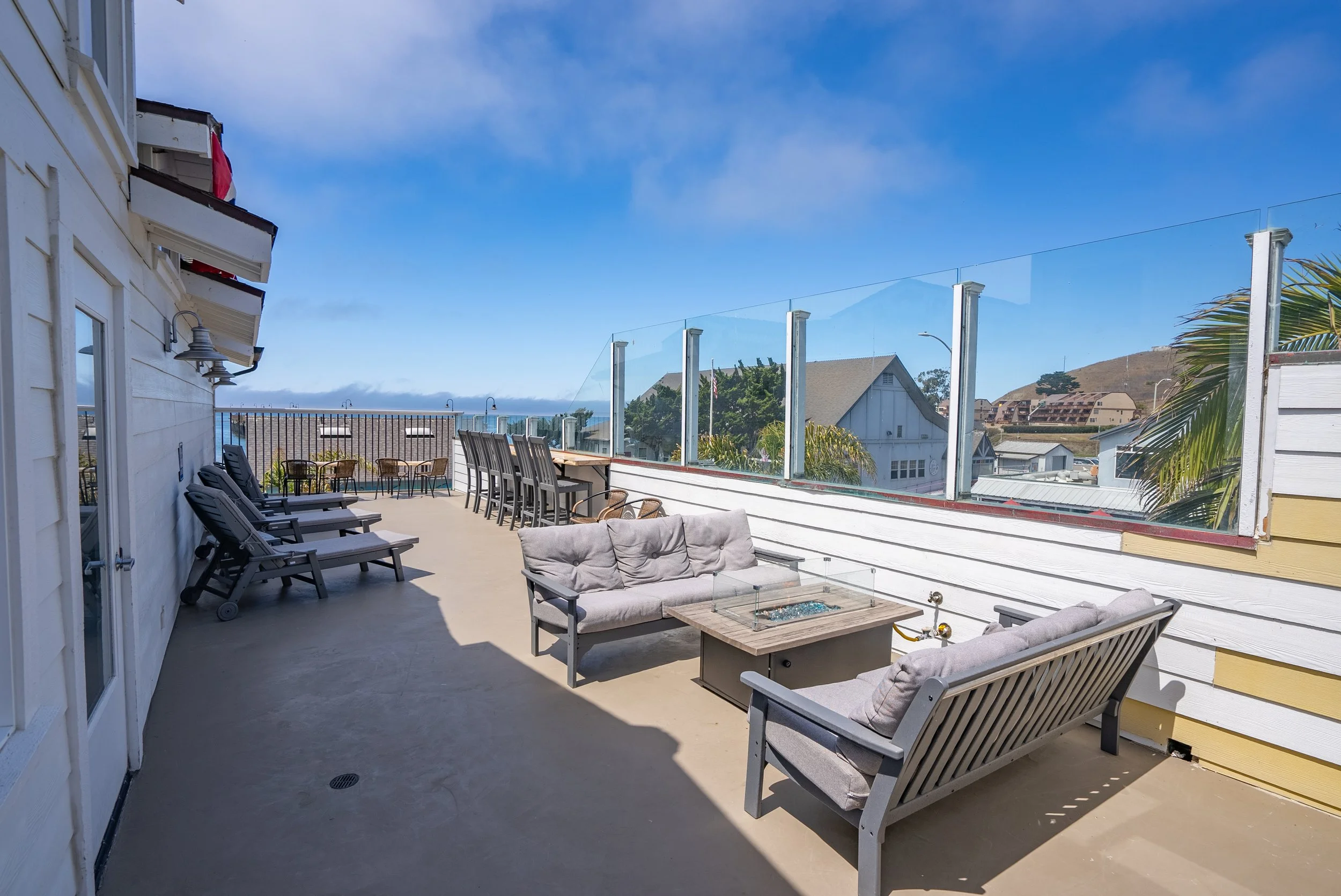 Outdoor rooftop patio area with lounge chairs, a sofa set, a coffee table, high-top chairs, and a glass railing overlooking neighboring houses and a hillside under a partly cloudy sky.