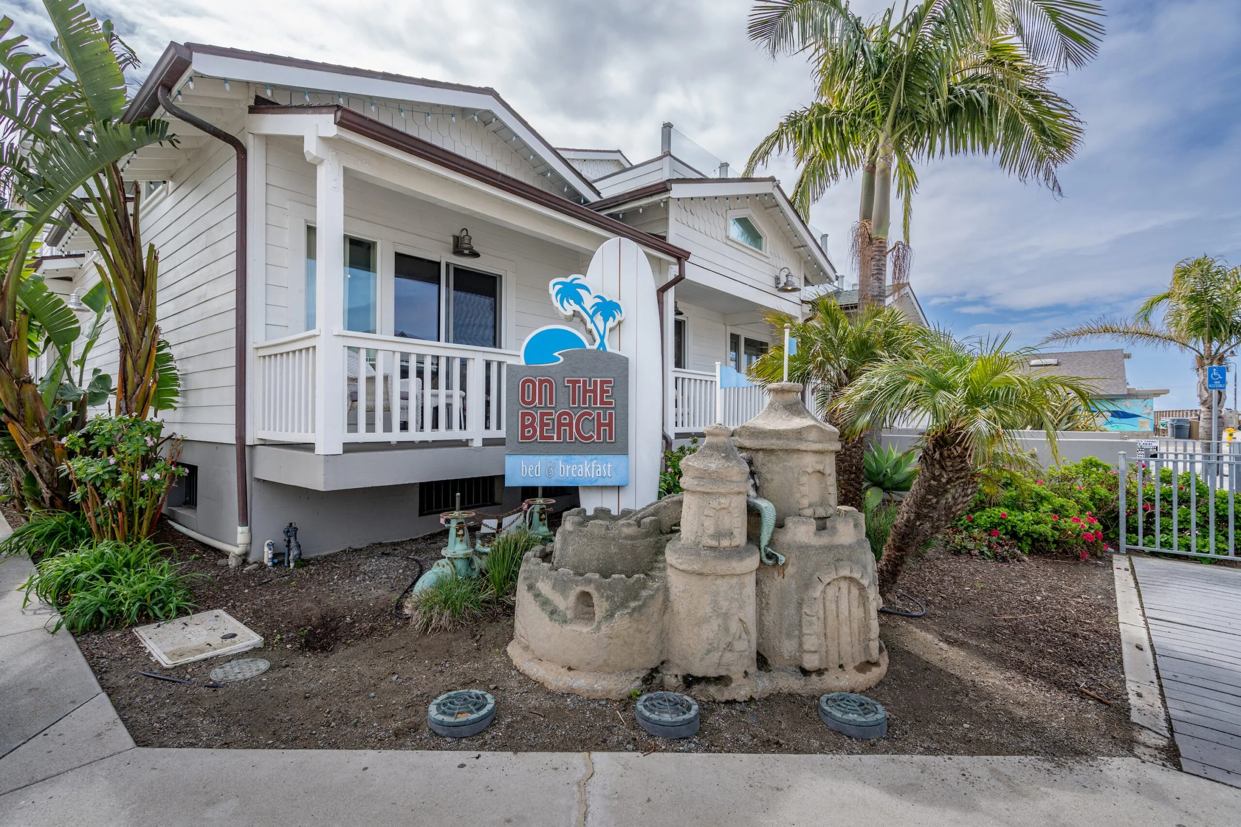 Beach-themed bed and breakfast with a white wooden building, a sign reading "On The Beach bed & breakfast," sandcastle garden decoration, palm trees, and a cloudy sky.