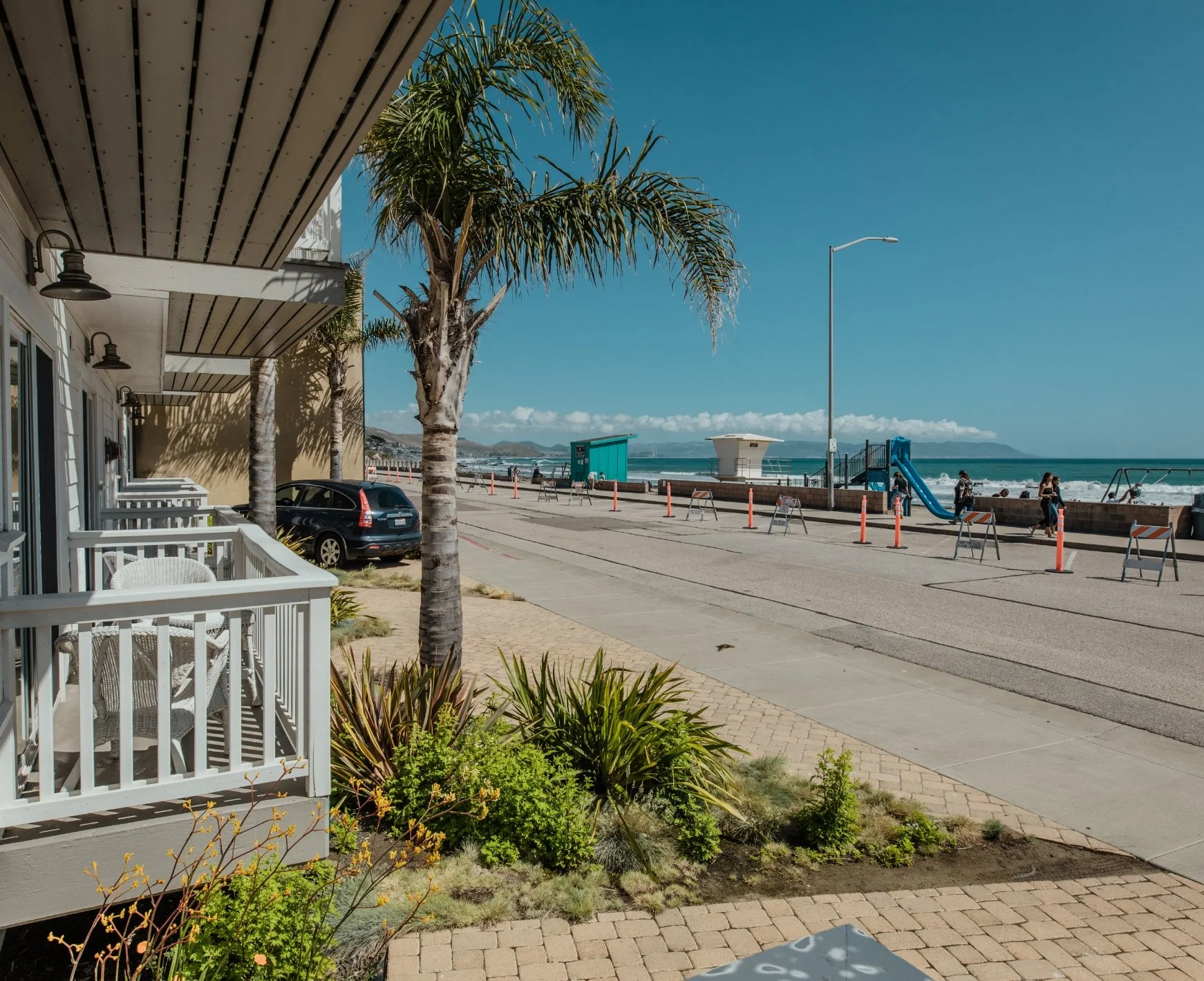 A sunny day at a beachside promenade with palm trees, a building with a porch, parked car, and a playground with a slide near the ocean, with people walking and a clear blue sky.