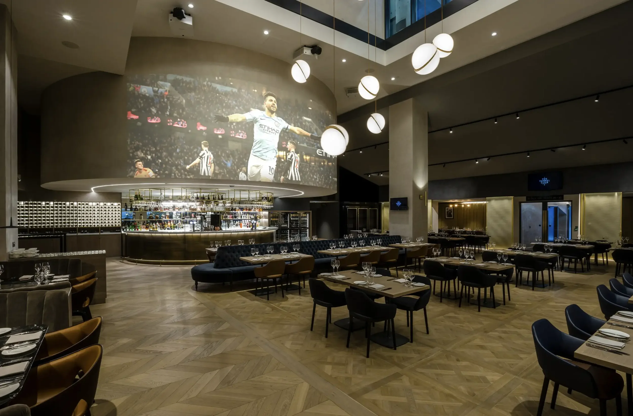 Manchester City Tunnel Club dining space with large-scale projection wall, globe lighting and parquet flooring — interior design by Studio Hayes.