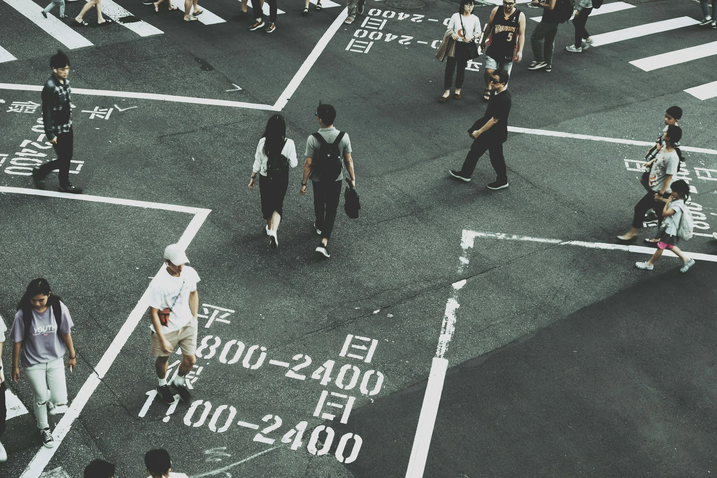 People crossing at an intersection on a Taipei city street, with white markings on the asphalt indicating instructions in the Chinese language. The scene is bustling with pedestrians, including men, women, and children.