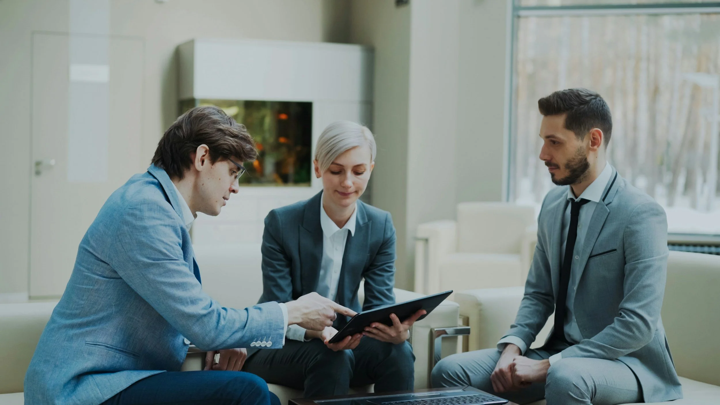 Three business professionals sitting on a beige sofa in a modern office, looking at a tablet device.