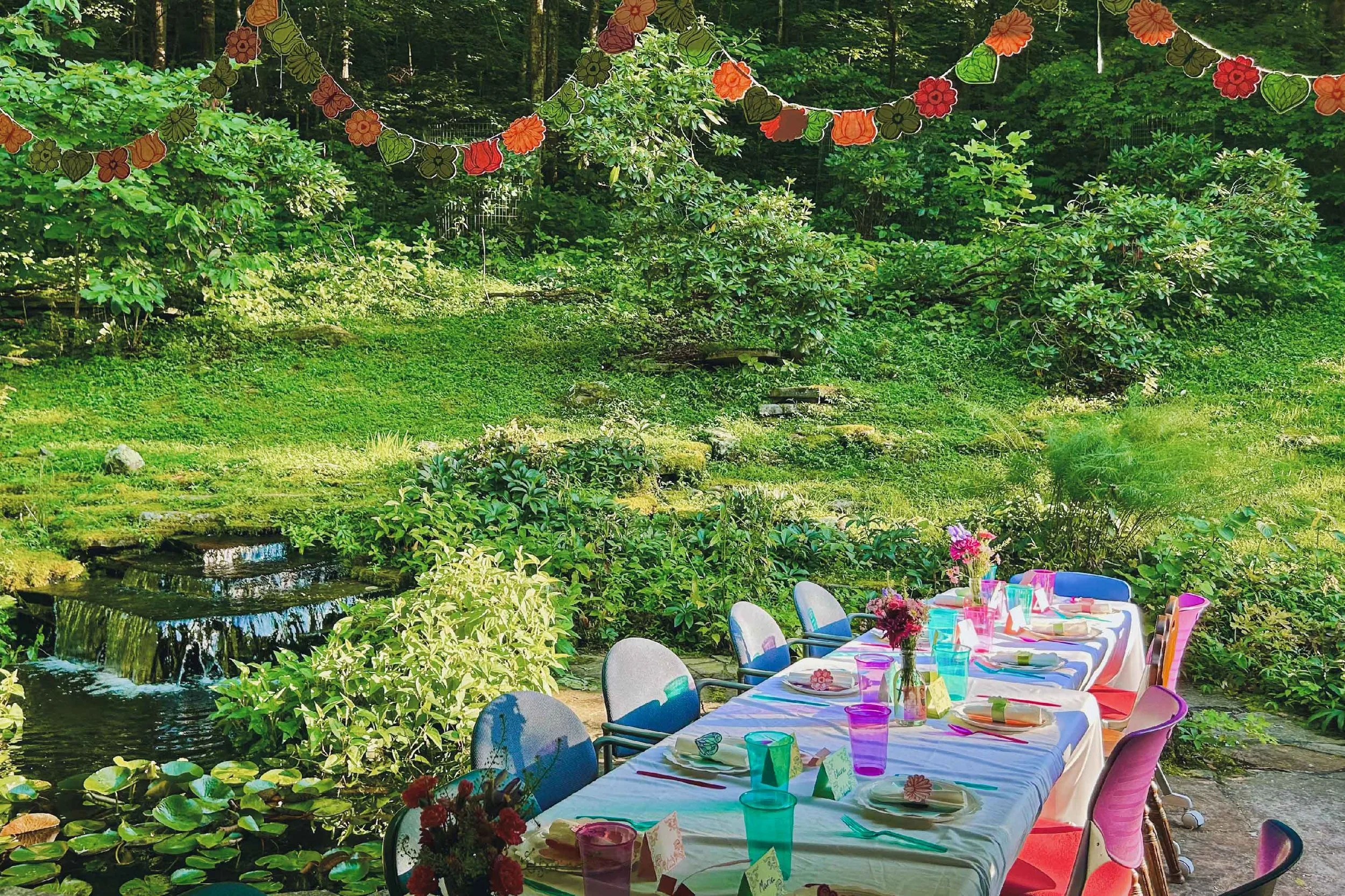 A long outdoor table set for a gathering, with colorful chairs, flower arrangements, and place settings, arranged in a lush garden near a small water feature, with decorative hanging flowers overhead.