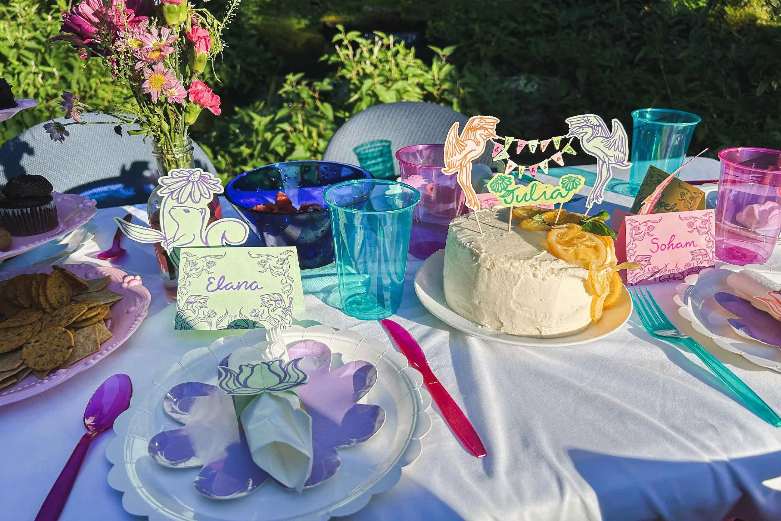 A colorful outdoor birthday party table with a white cake, decorative place cards, a flower arrangement, and various snacks, set on a white tablecloth surrounded by chairs and greenery.