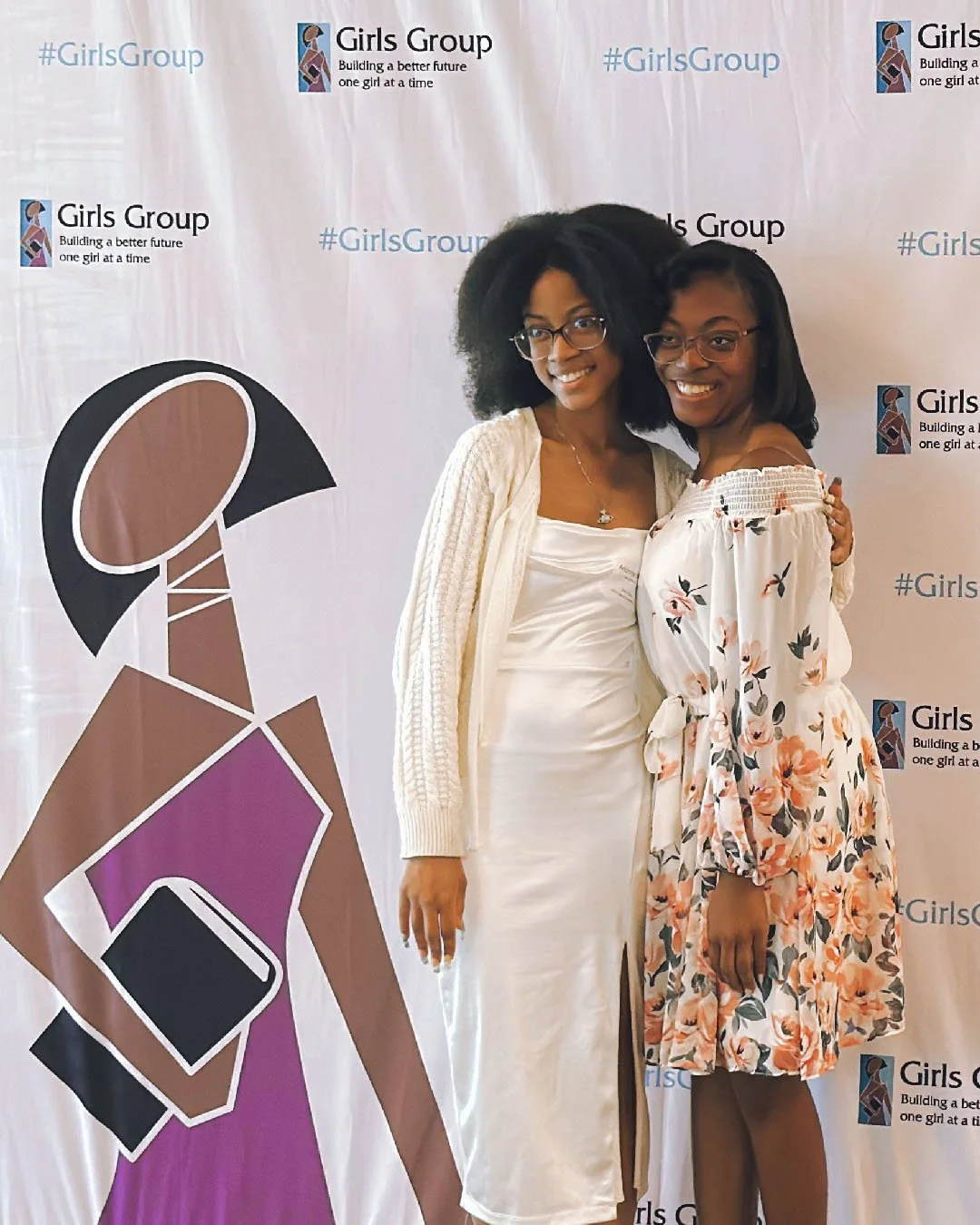 Two women smiling and hugging in front of a Girls Group backdrop with a cartoon logo of a girl with glasses and text that says, 'Building a better future one girl at a time.'