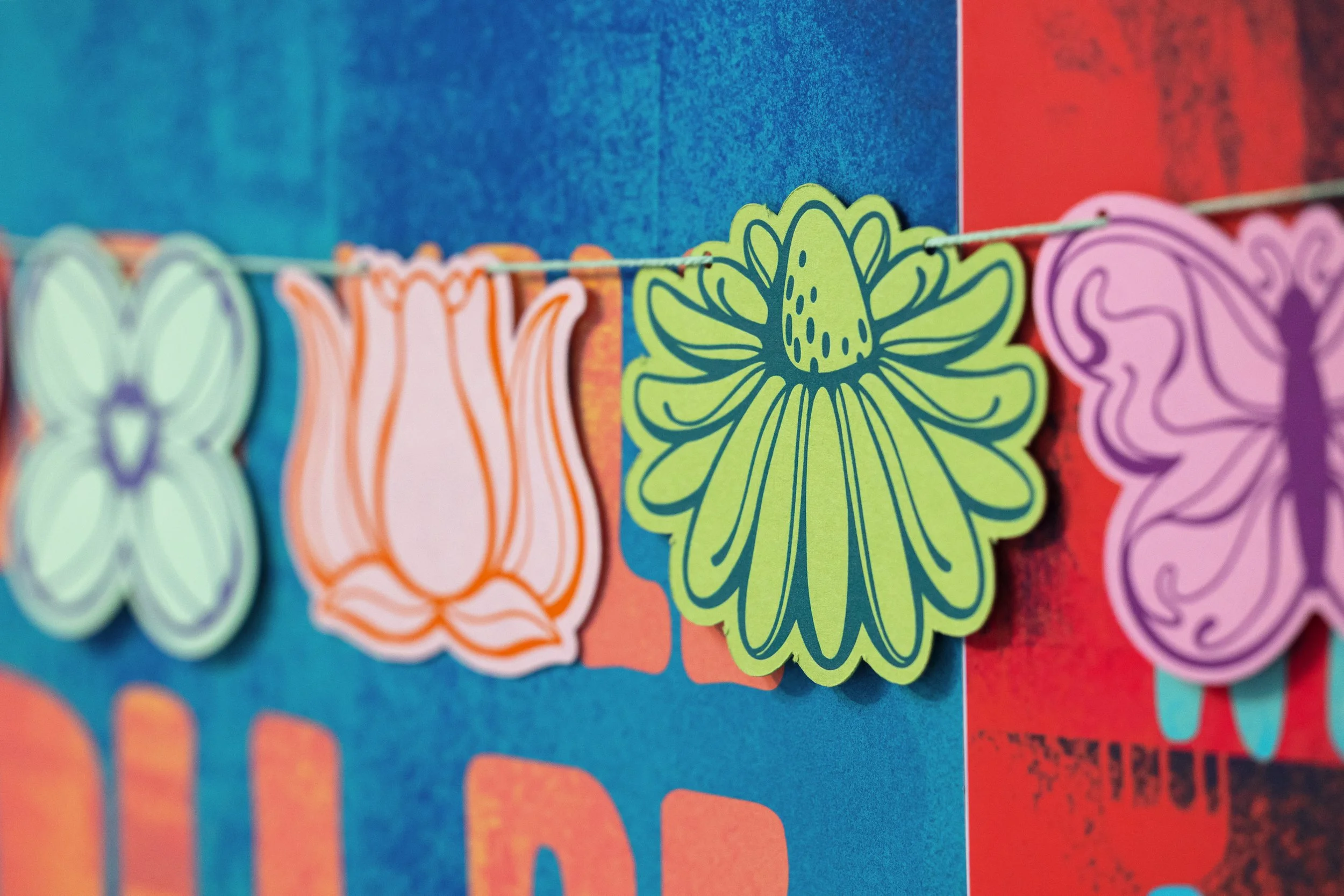 Colorful paper cutouts of flowers, butterflies, and insects hanging on a string against a vibrant blue and red background.