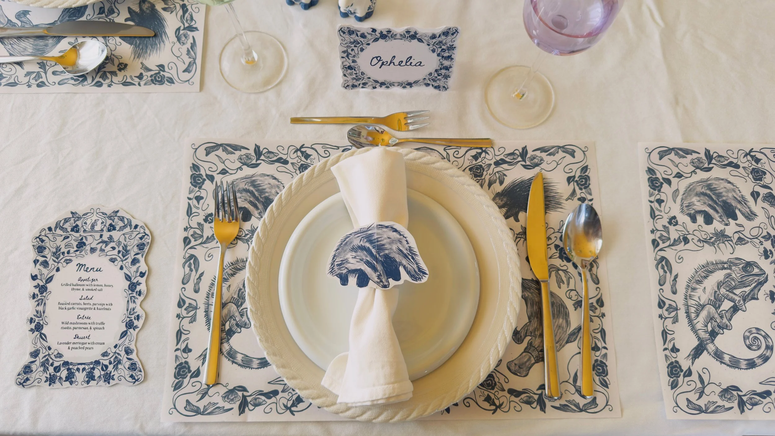 A table setting with a white plate, a beige napkin with a blue and white feather design, yellow and silver cutlery, and a blue and white themed tablecloth with animals and floral patterns. There is a name card labeled 'Ophelia' and a menu with a simi