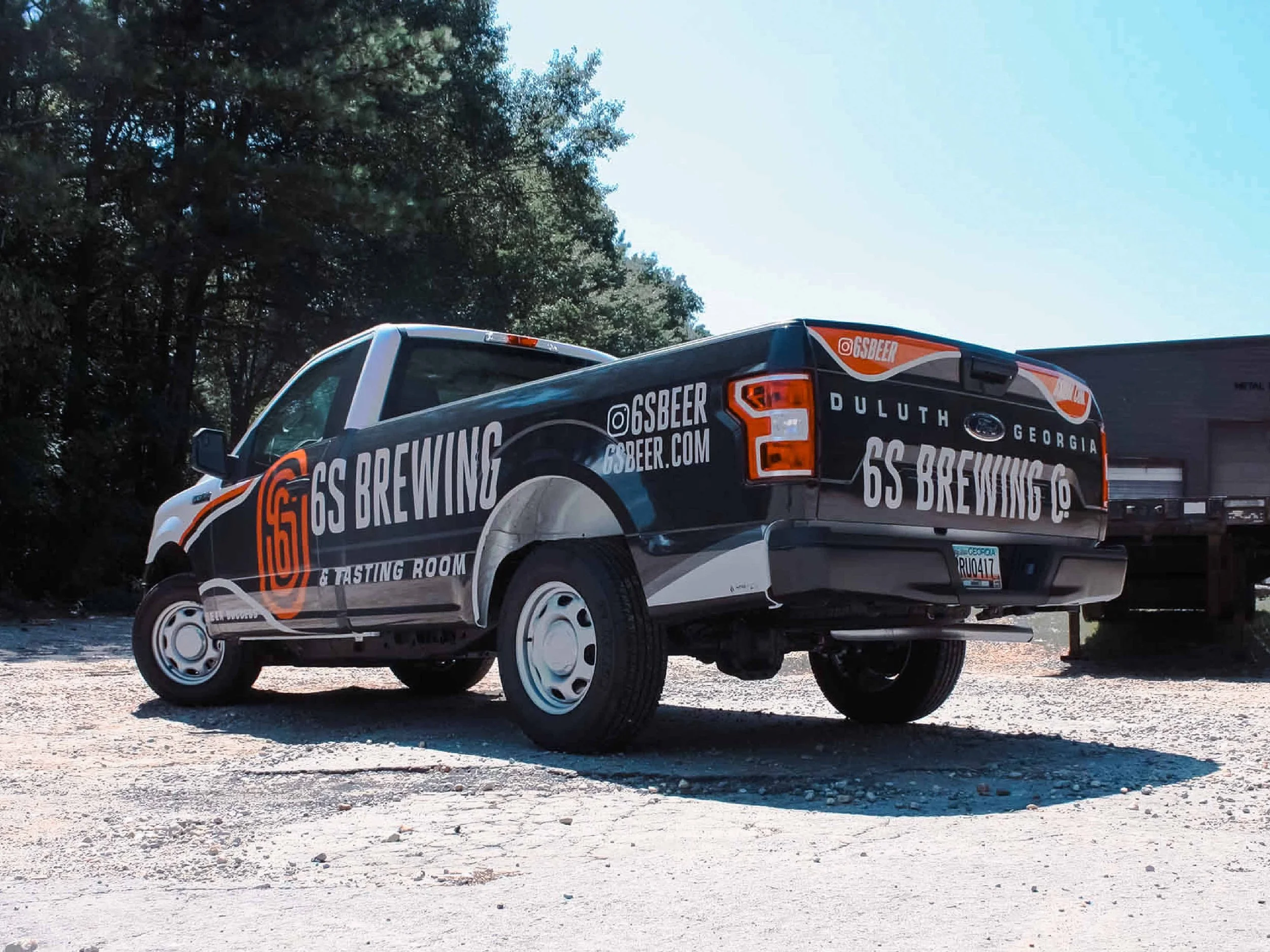 A pickup truck with advertising for 6S Brewing on its sides, parked outdoors on a gravel surface with trees and a trailer in the background.