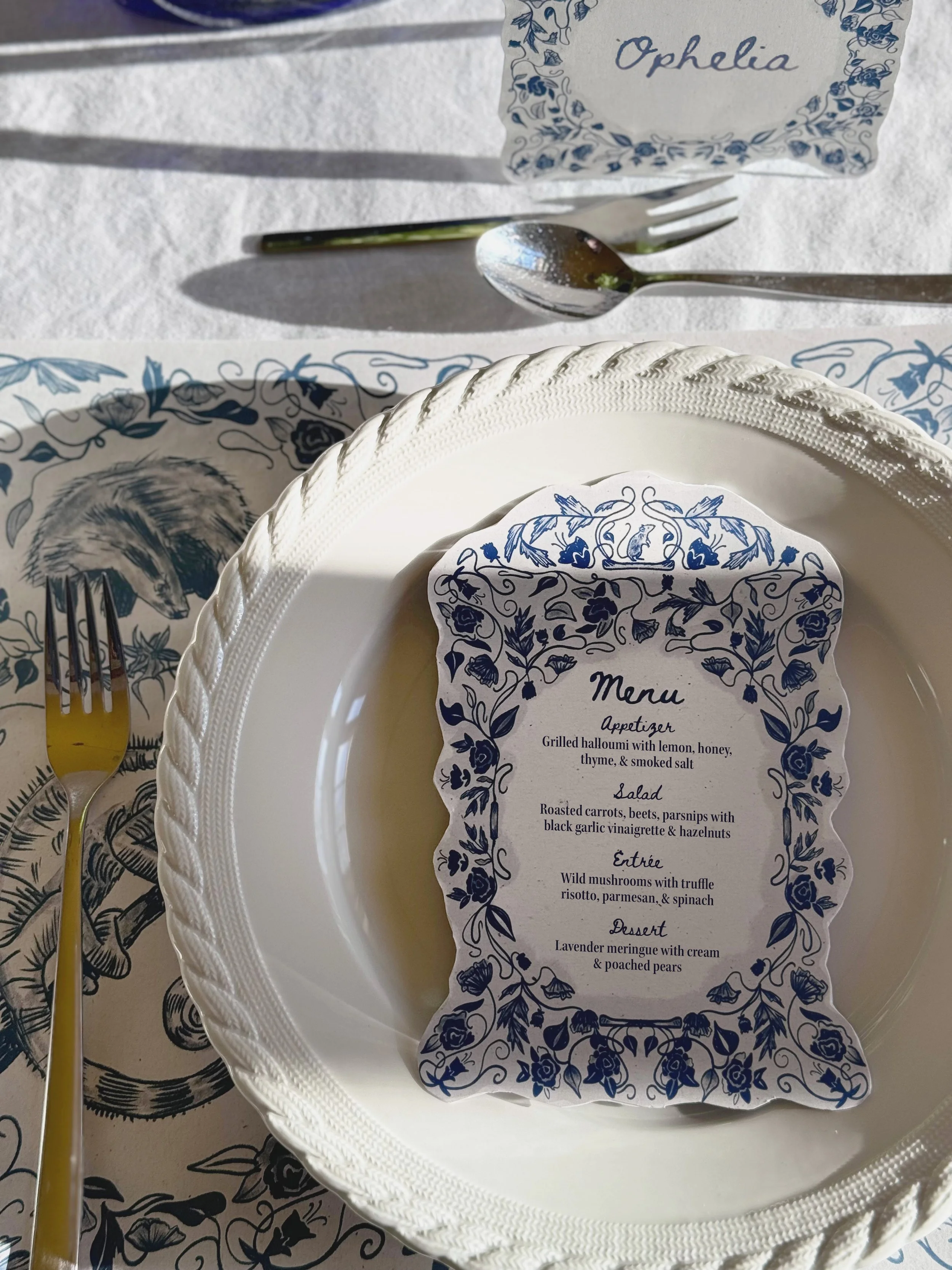 A table setting with a white plate, a knife and fork, a silver spoon, and a menu with a blue floral border. The menu includes appetizer, salad, entrée, and dessert items, and the tablecloth and placemat feature a matching blue floral design.