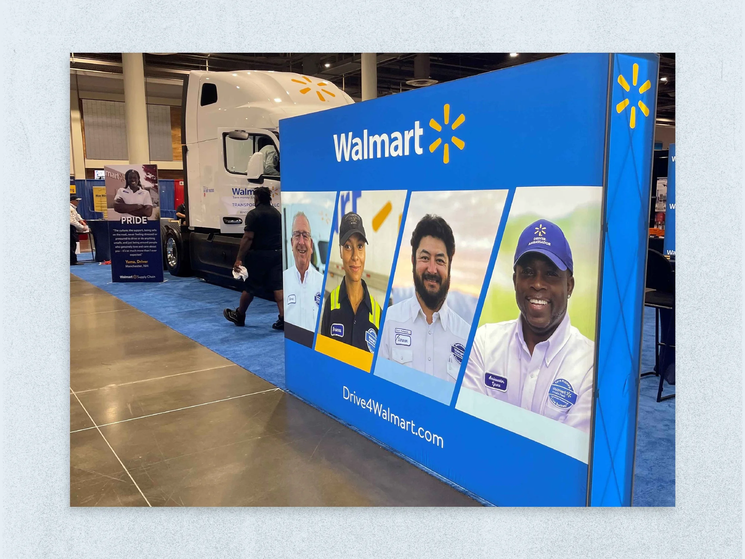 Walmart promotional display with large banner featuring four Walmart employees and a large truck in the background, displayed at an indoor event or expo.