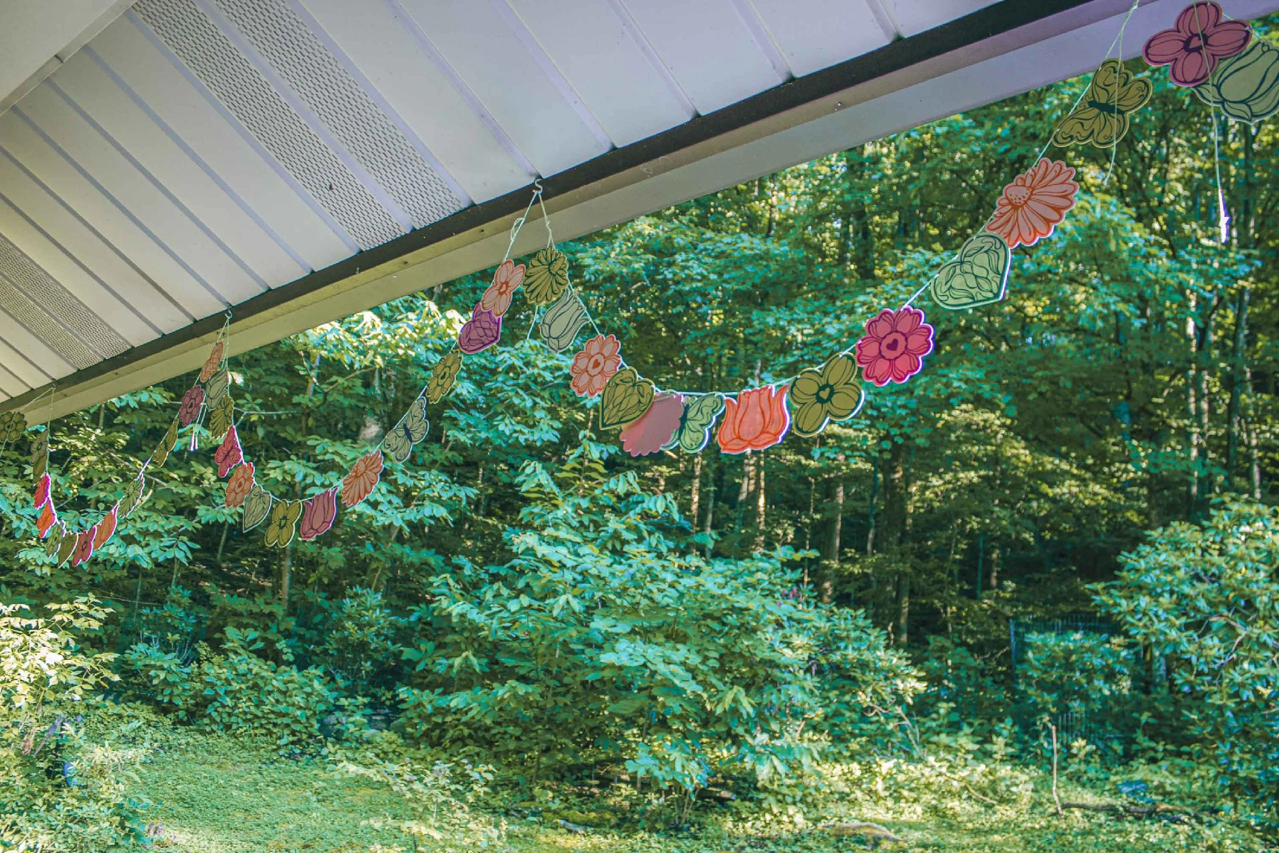 Colorful paper flower garlands hanging under a porch roof, with a background of green trees and bushes.