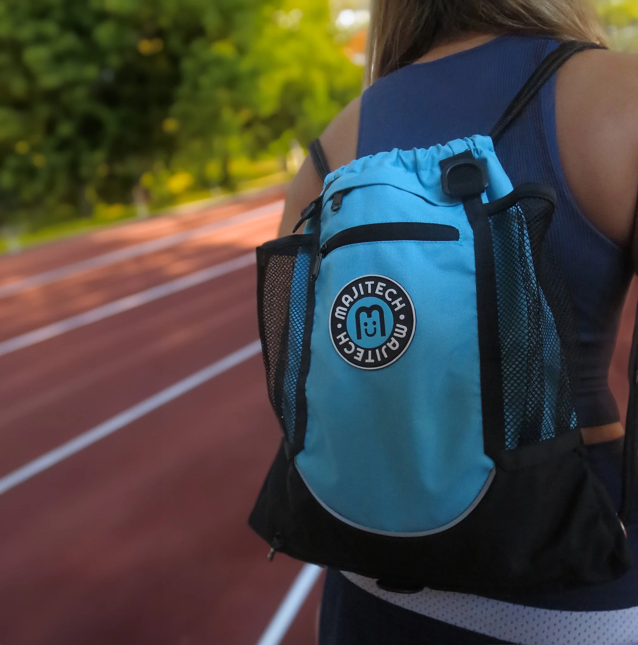 A person wearing a blue tank top and a black sports bra with a small blue and black backpack standing on a running track with green trees in the background.