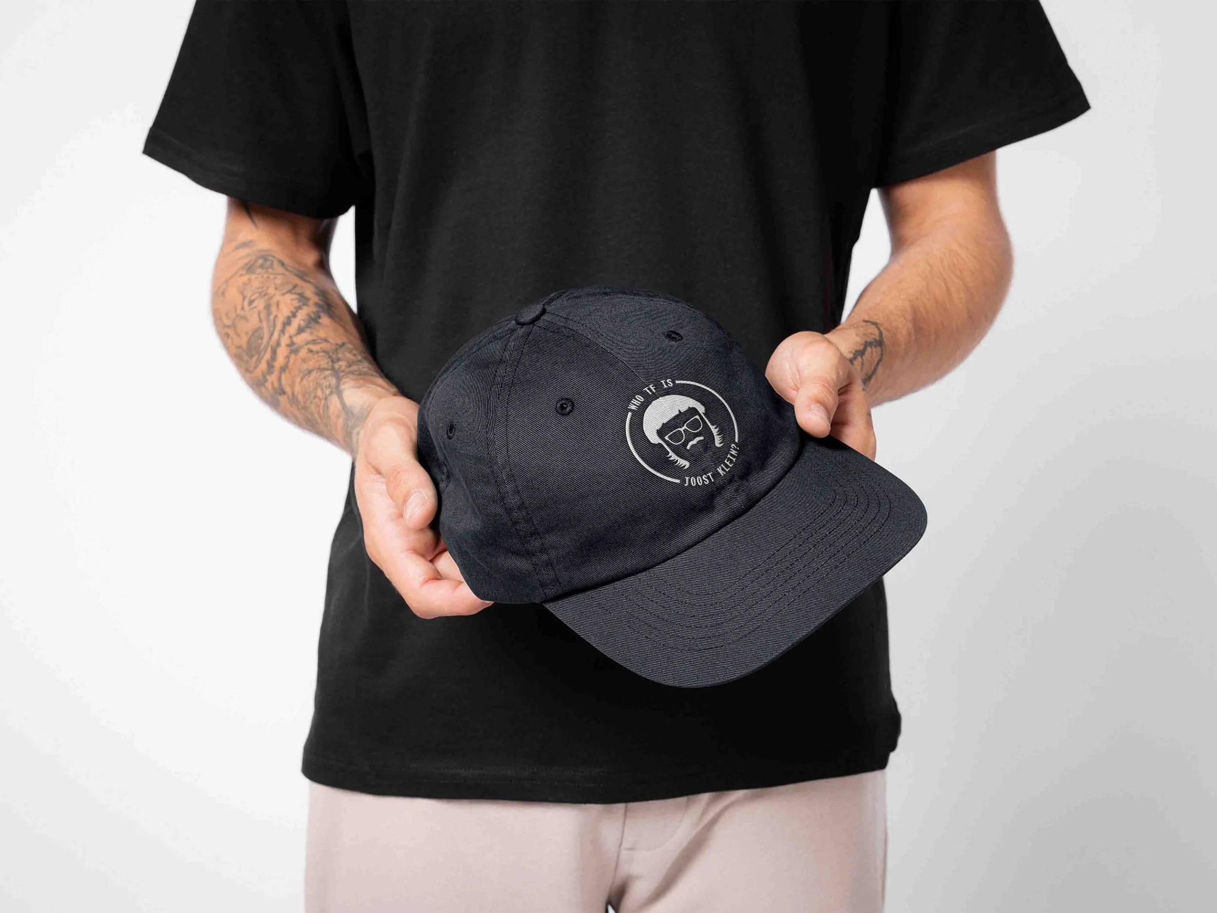 Person holding a black cap with white embroidered logo and text, wearing a black t-shirt and having tattooed arms, against a plain white background.