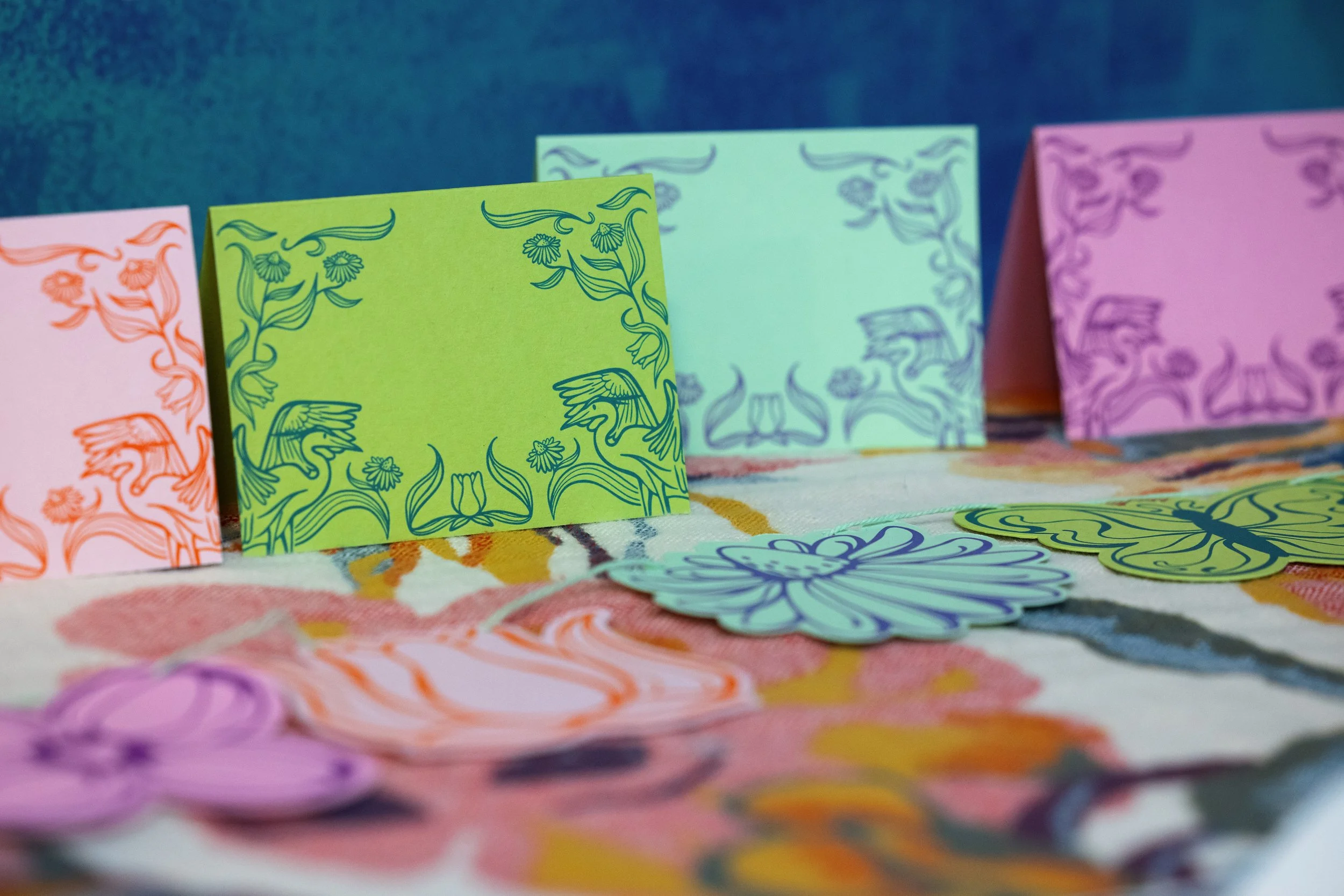 Colorful paper crafts with intricate line drawings of birds, flowers, and floral patterns in pink, green, blue, and purple, arranged on a table with a colorful cloth.