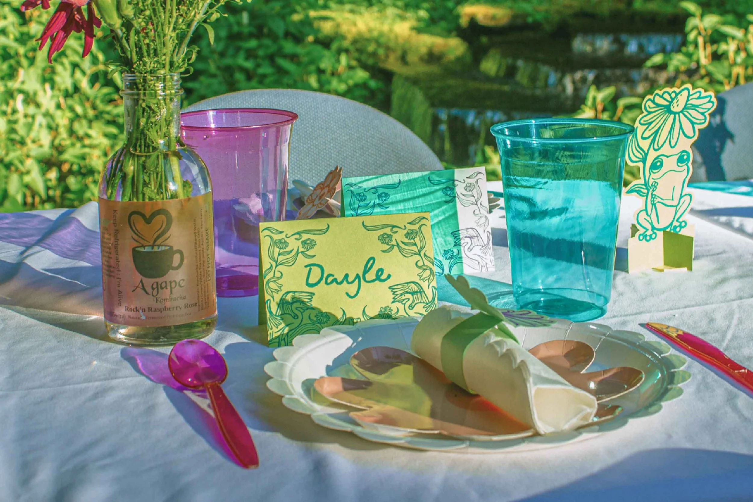 Outdoor table set with colorful glassware, the restaurant's name tag, decorative paper cutouts, a rolled napkin, and a plate of food, with a background of green foliage.