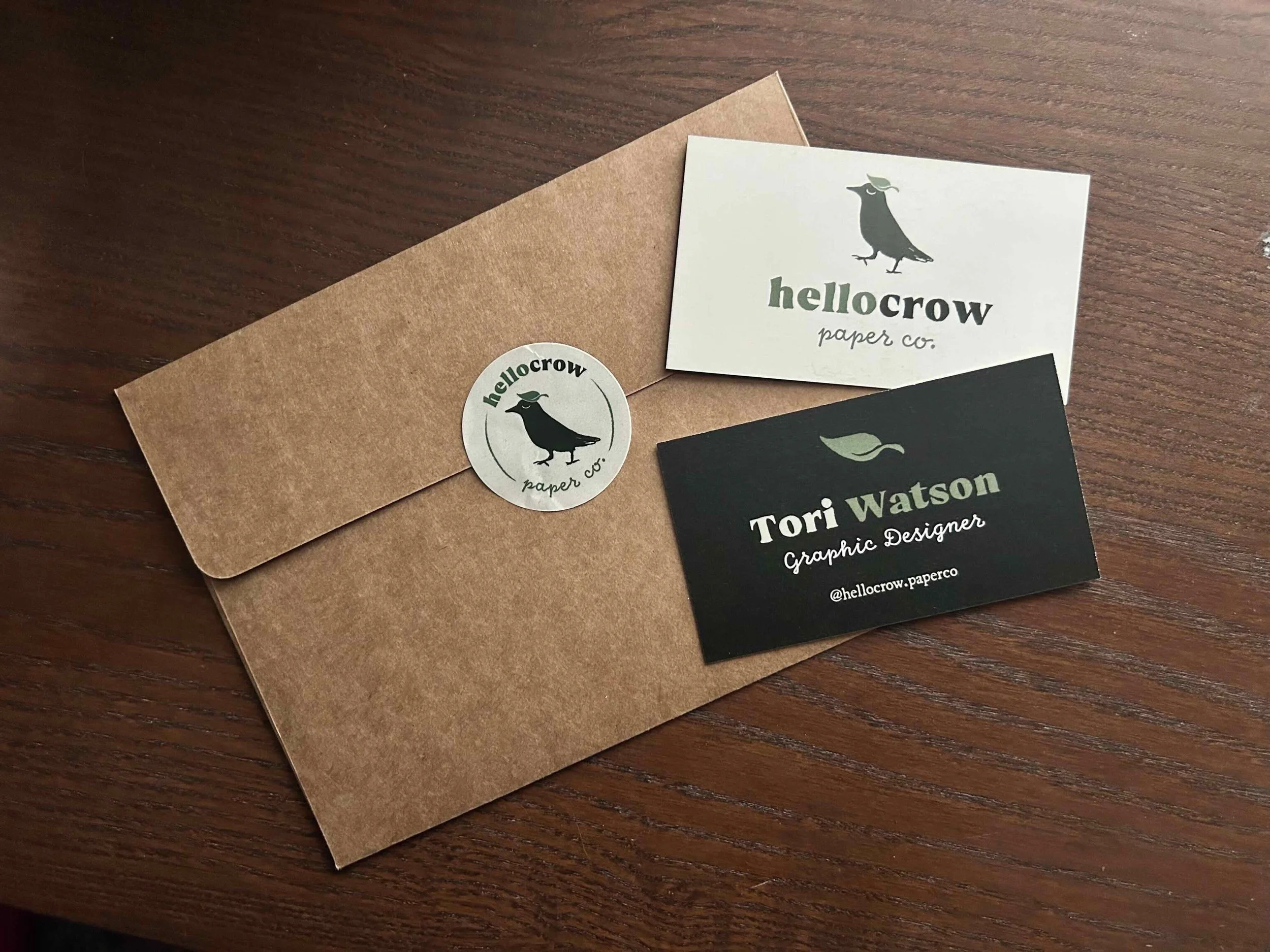 A brown paper envelope with a Hello Crow Paper Co. sticker, a white business card with a crow logo and the company name, and a black business card with the name Tori Watson, a graphic designer, and social media handle, all placed on a dark wooden sur