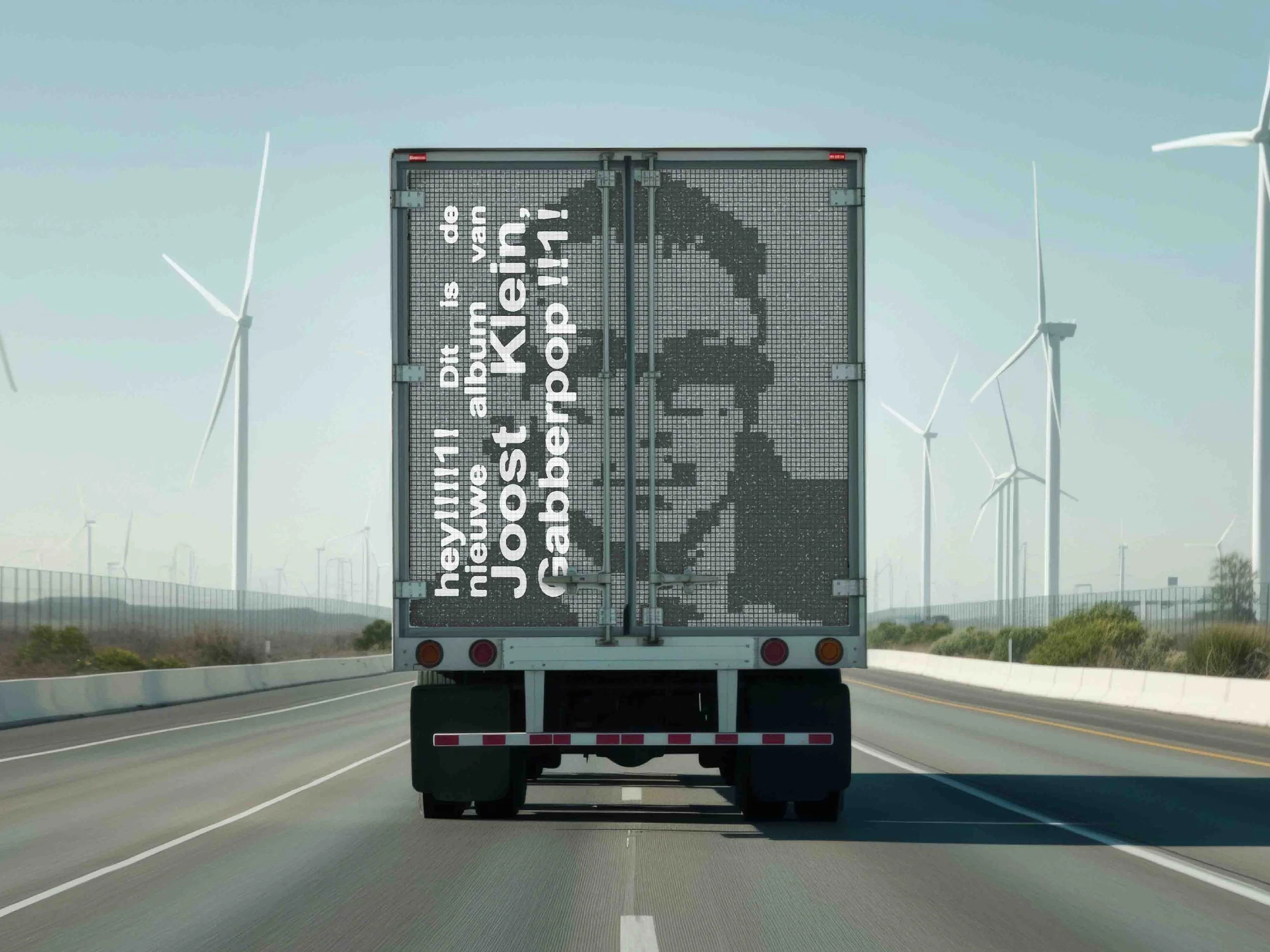 A truck on a highway with a digital display on the back showing a pixelated image of a person and text that reads, "hey! nick, we just album de van. joost abakert popin!" Wind turbines are visible in the background.