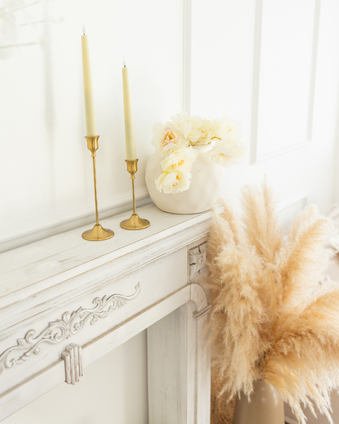Vases de fleurs blanches sur une cheminée blanche décorée, avec deux bougeoirs dorés et de la pampa beige à proximité.