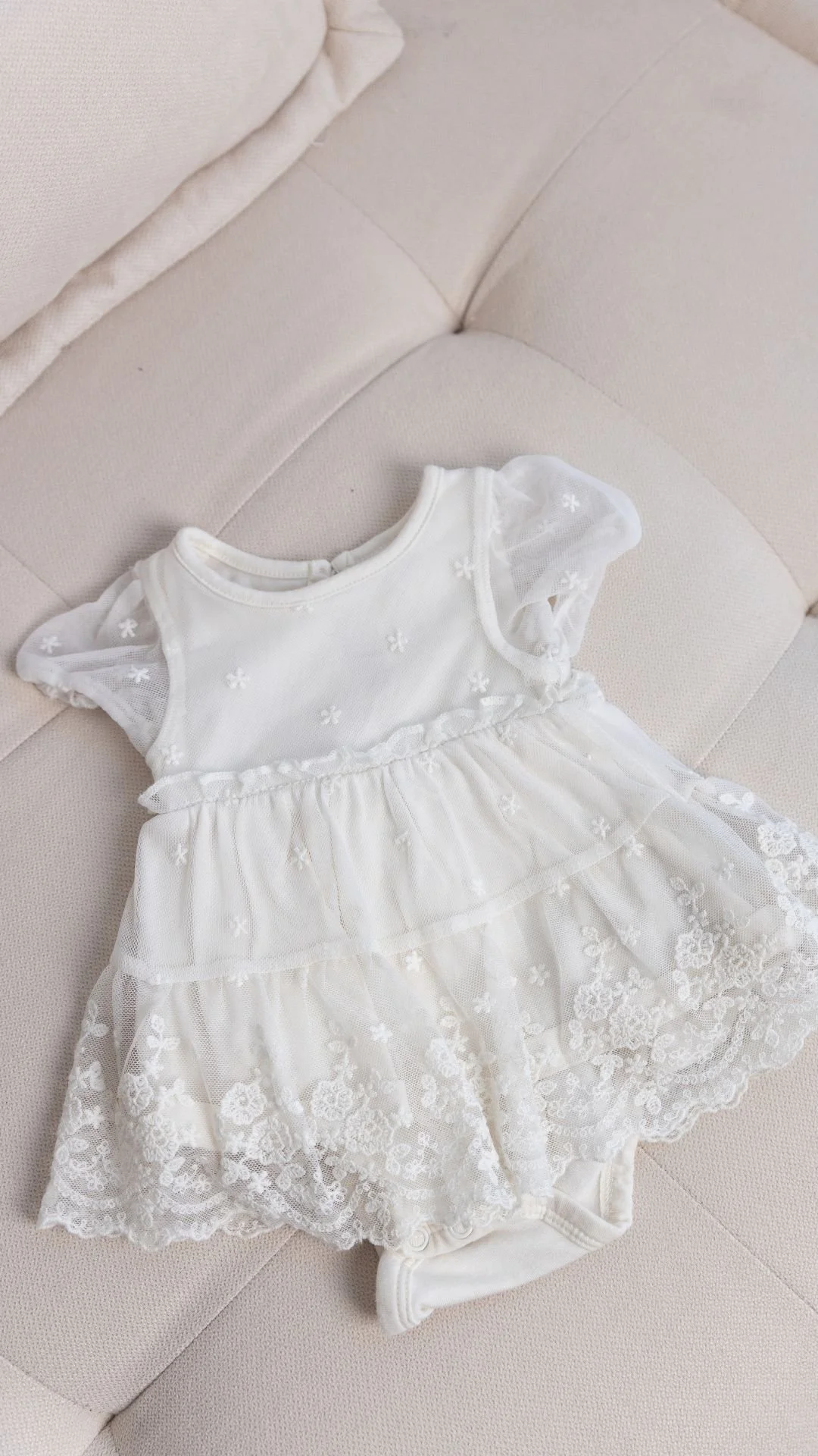 Une petite robe blanche avec des manches en tulle et des broderies florales, posée sur un canapé beige.