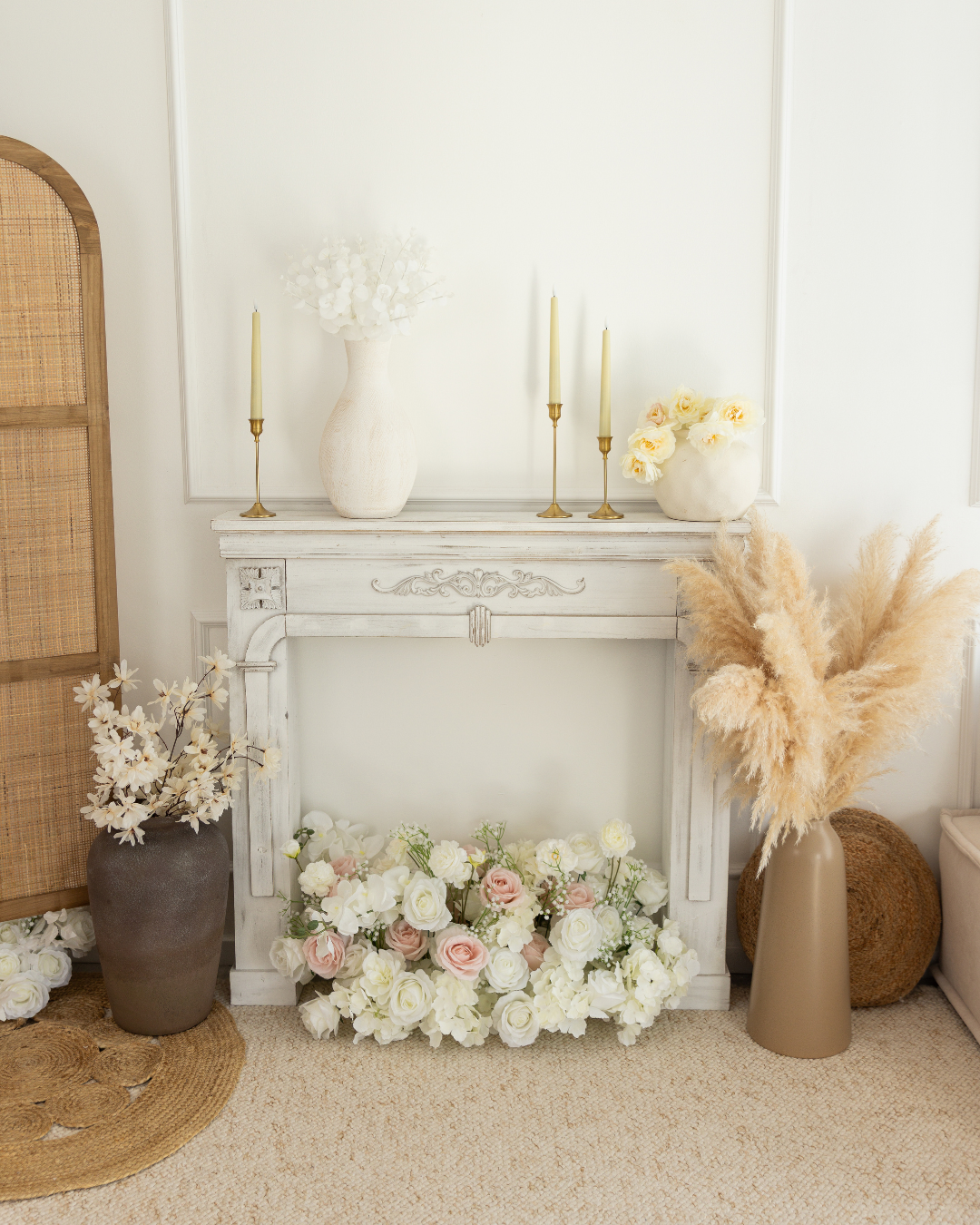 Décoration intérieure avec une cheminée blanche, vase blanc avec fleurs blanches, bougies dorées, bouquet de fleurs roses et blanches, et pampas dans un vase beige.