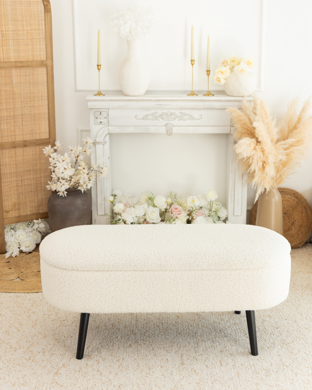 Un panier beige dans une pièce décorée avec un manteau de cheminée blanc, des vases avec des fleurs blanches, des bougies et des plumes