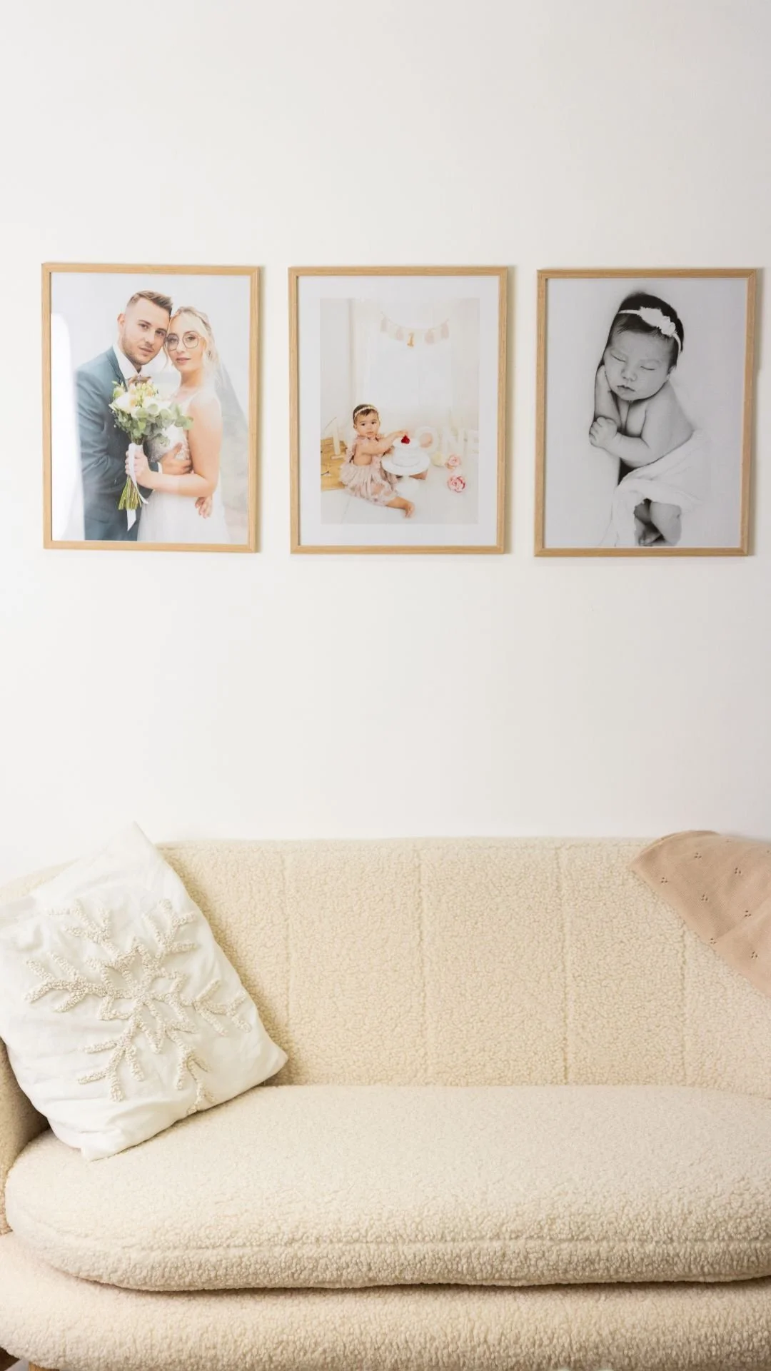Trois cadres photos accrochés au mur, dont un portrait d'un couple en mariage, un de bébé avec un gâteau, et un d'une petite fille endormie, au-dessus d'un canapé beige avec un coussin blanc à motif flocon de neige.