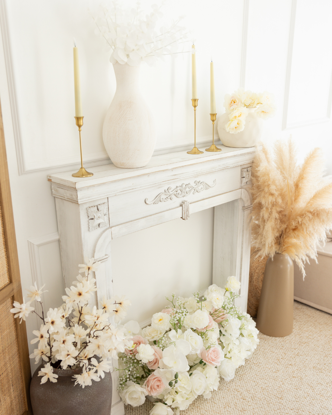 Décor intérieur avec une cheminée blanche, vases avec des fleurs blanches et roses, bougies dans des chandeliers en laiton, et un bouquet de pampas. Style pastel, lumineux et élégant.