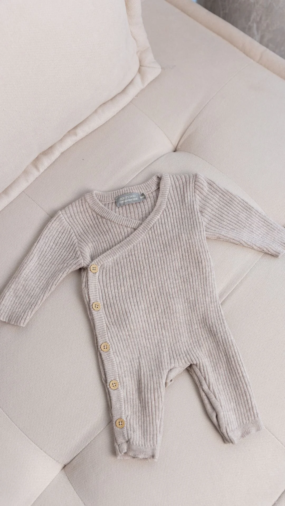 Une combinaison pour bébé en tricot rayé beige claire posée sur un canapé beige.