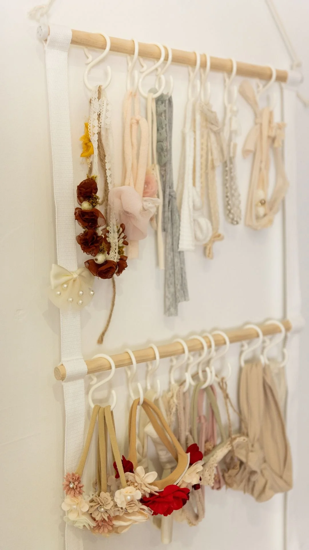 Une décoration murale composée de deux rangées de barres en bois avec des crochets suspendant divers accessoires en tissu, comme des bandeaux, des rubans et des accessoires de cheveux, dans des tons pastel et neutres.