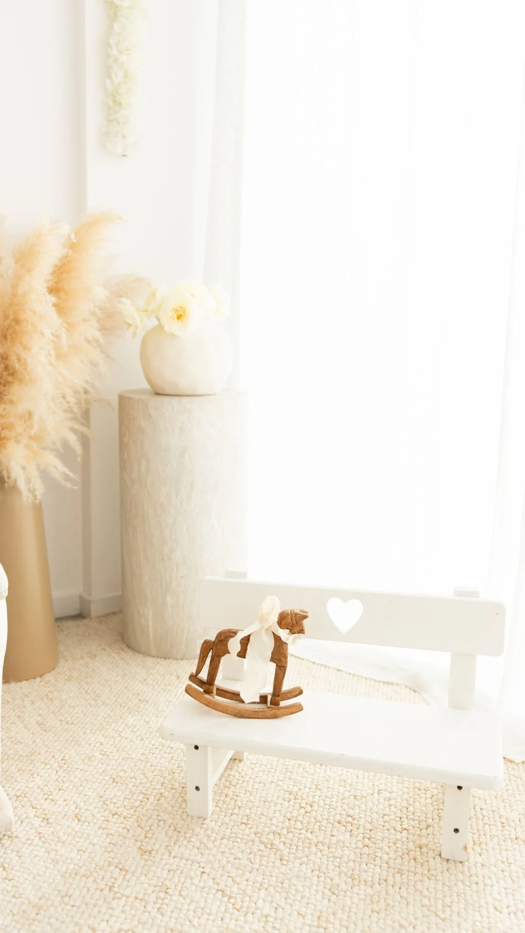 Un petit banc blanc avec un cœur découpé dans le dossier, une petite poupée en bois de cheval à bascules posée dessus, dans une pièce décorée en style neutre avec une grande cascade de plumes dans un vase beige, une lampe cylindrique beige en arrière